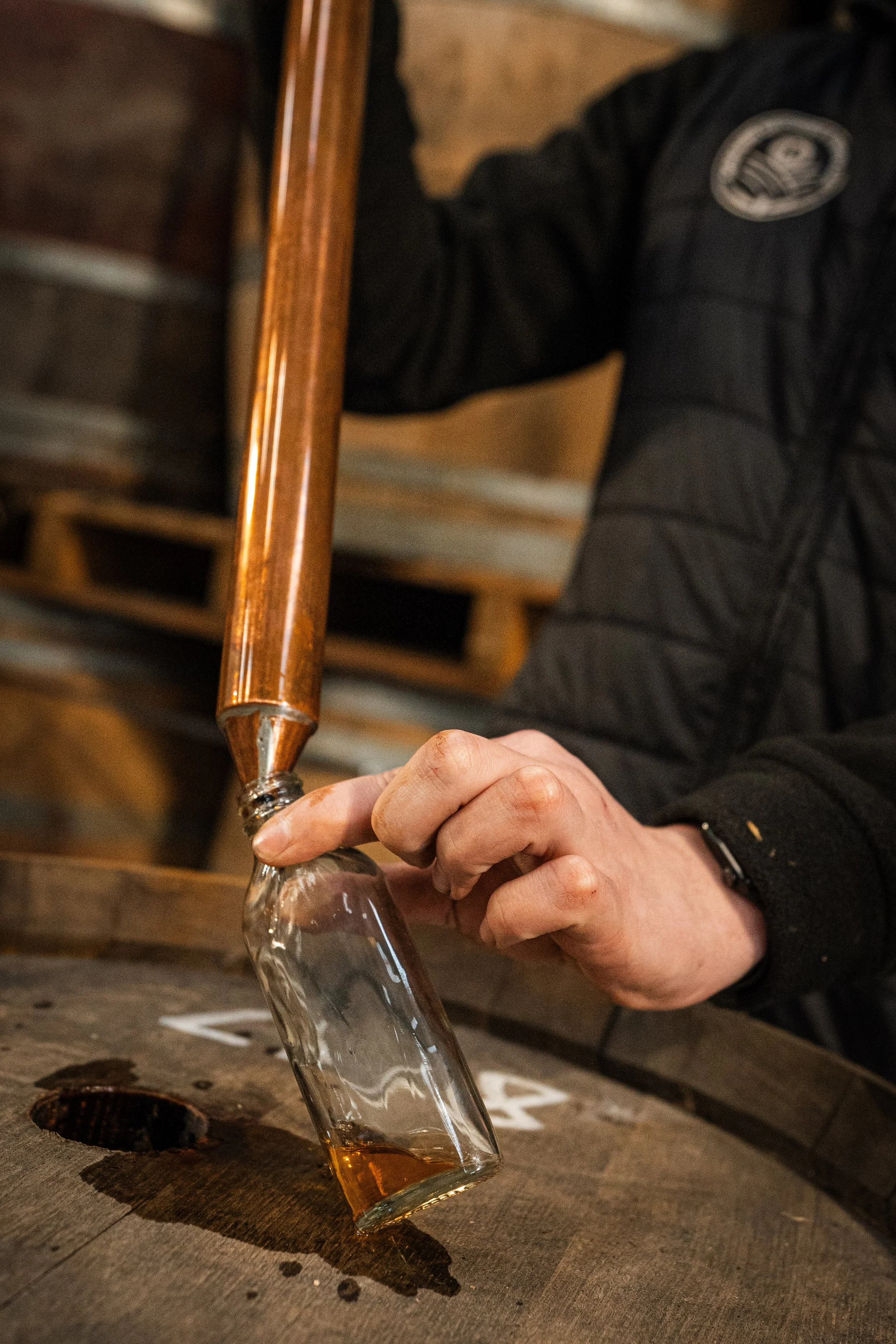 Person pouring whiskey from a copper still into a glass on a wooden barrel.