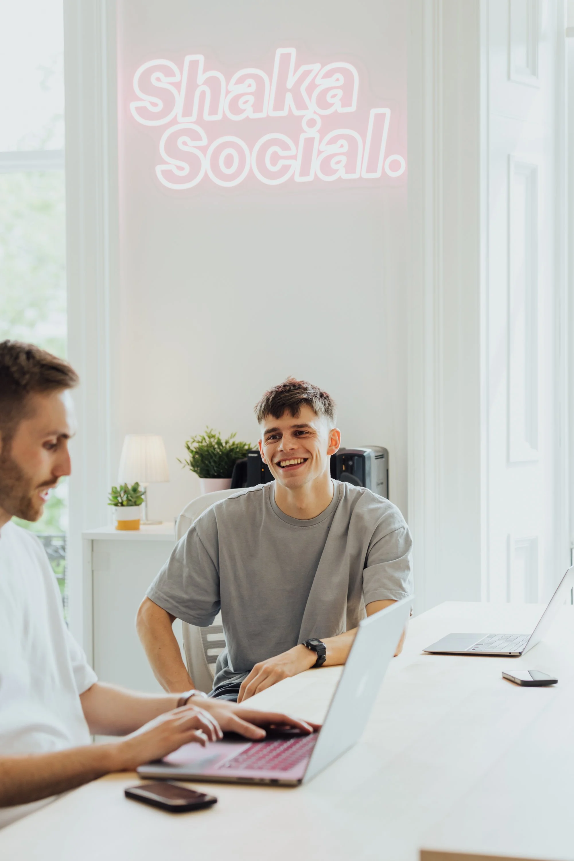 Two young men sitting at a white table in a bright room with large windows, smiling and talking. There are laptops, a smartphone, and a small plant on the table. A neon sign on the wall reads "Shaka Social." in the background.