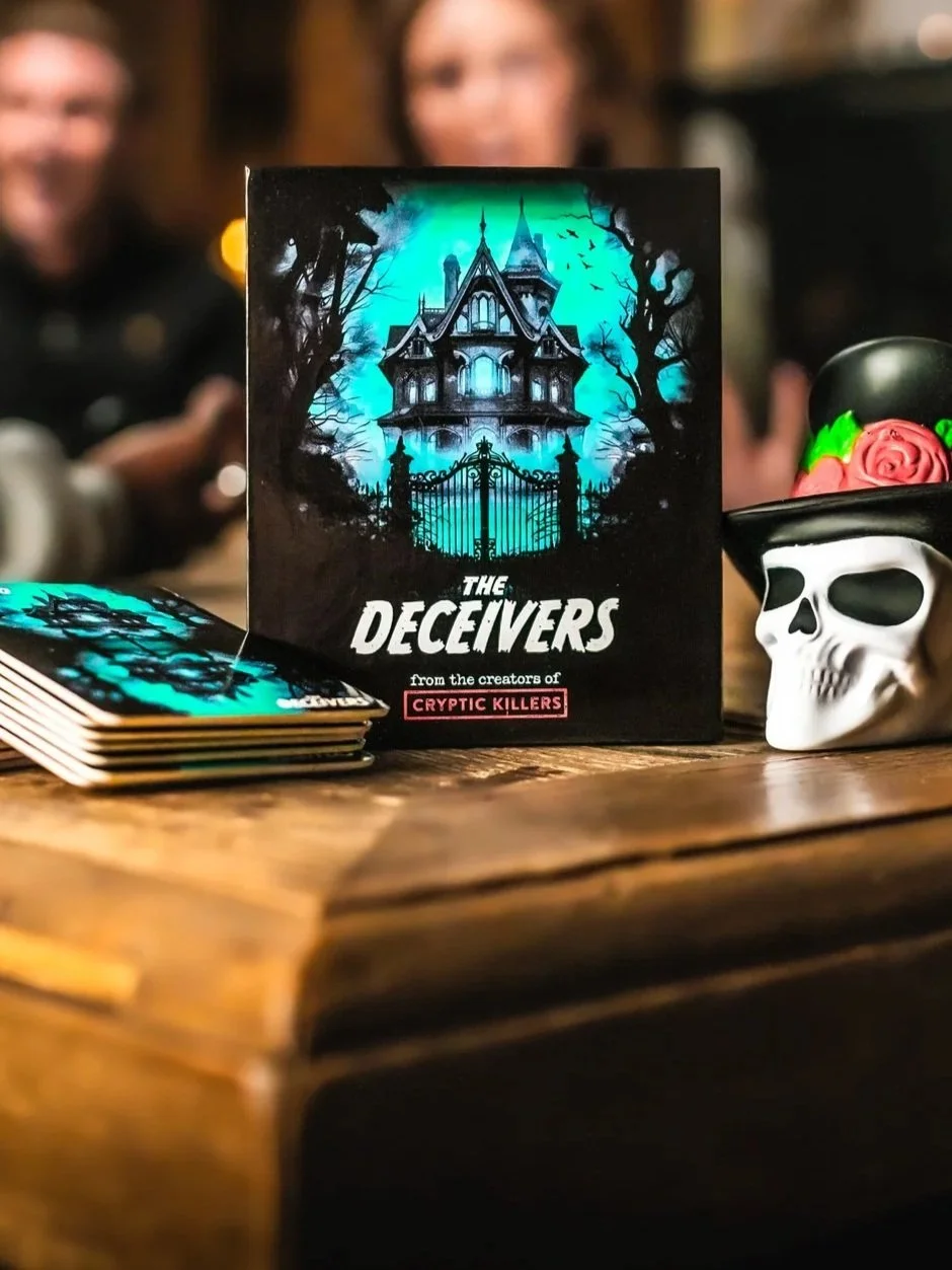 A spooky haunted house illustration on the box of the card game 'The Deceivers' placed on a wooden table, with a decorative skull wearing a black top hat adorned with pink roses and green accents, and a blurred background showing two people.