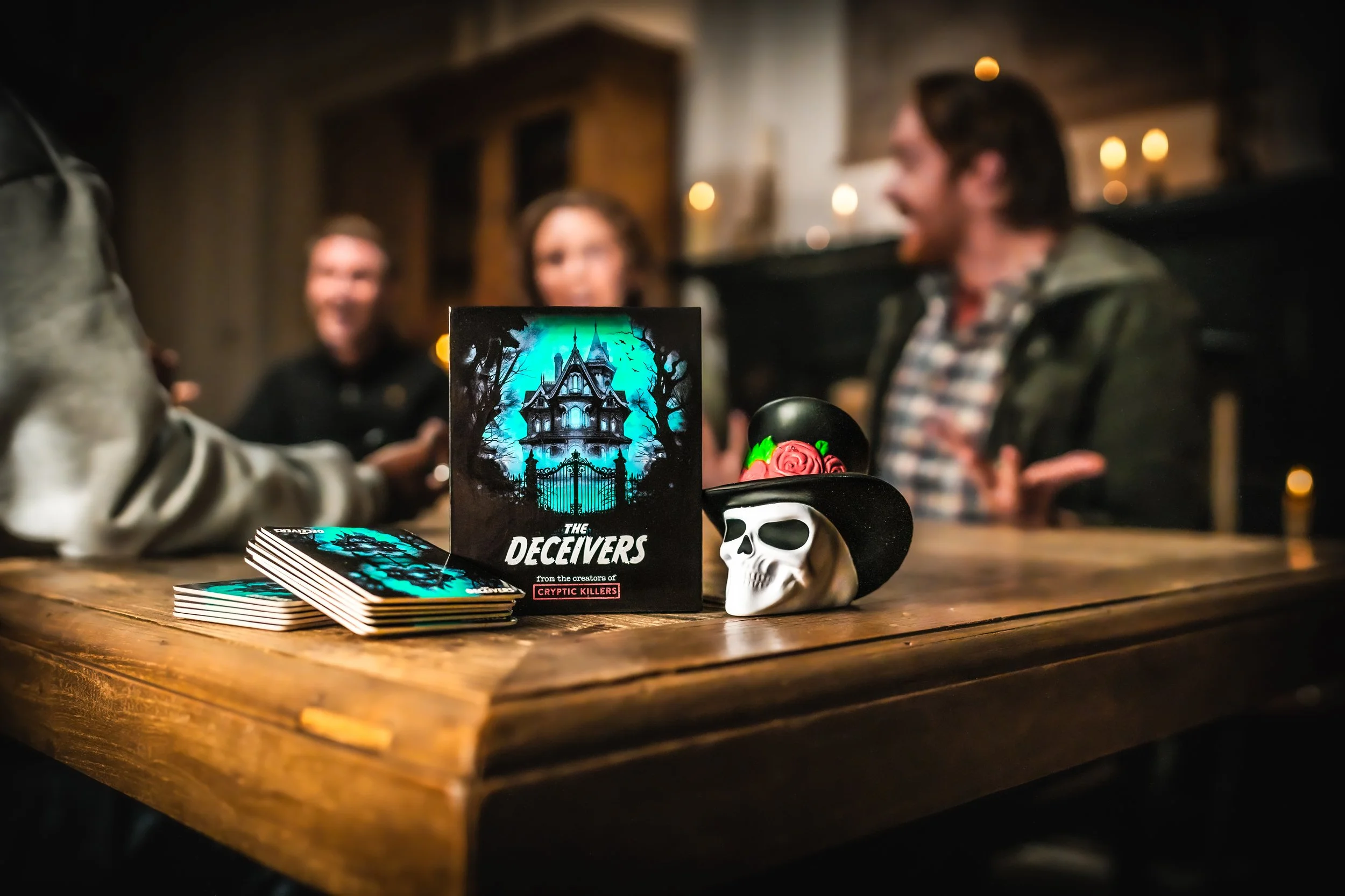 A tabletop with a horror-themed game called 'The Deceivers', featuring a house on the cover with haunted trees. There is a deck of cards and a skull with a black hat decorated with a pink rose and green leaves. In the background, three people are sitting at the table, talking and smiling in a cozy, dimly lit room with lit candles.