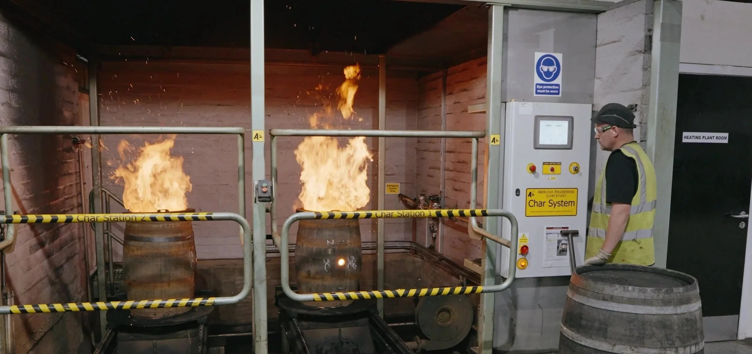 two barrels igniting flames inside whisky casks in a controlled environment, a safety officer observing, and caution tape indicating a designated station.