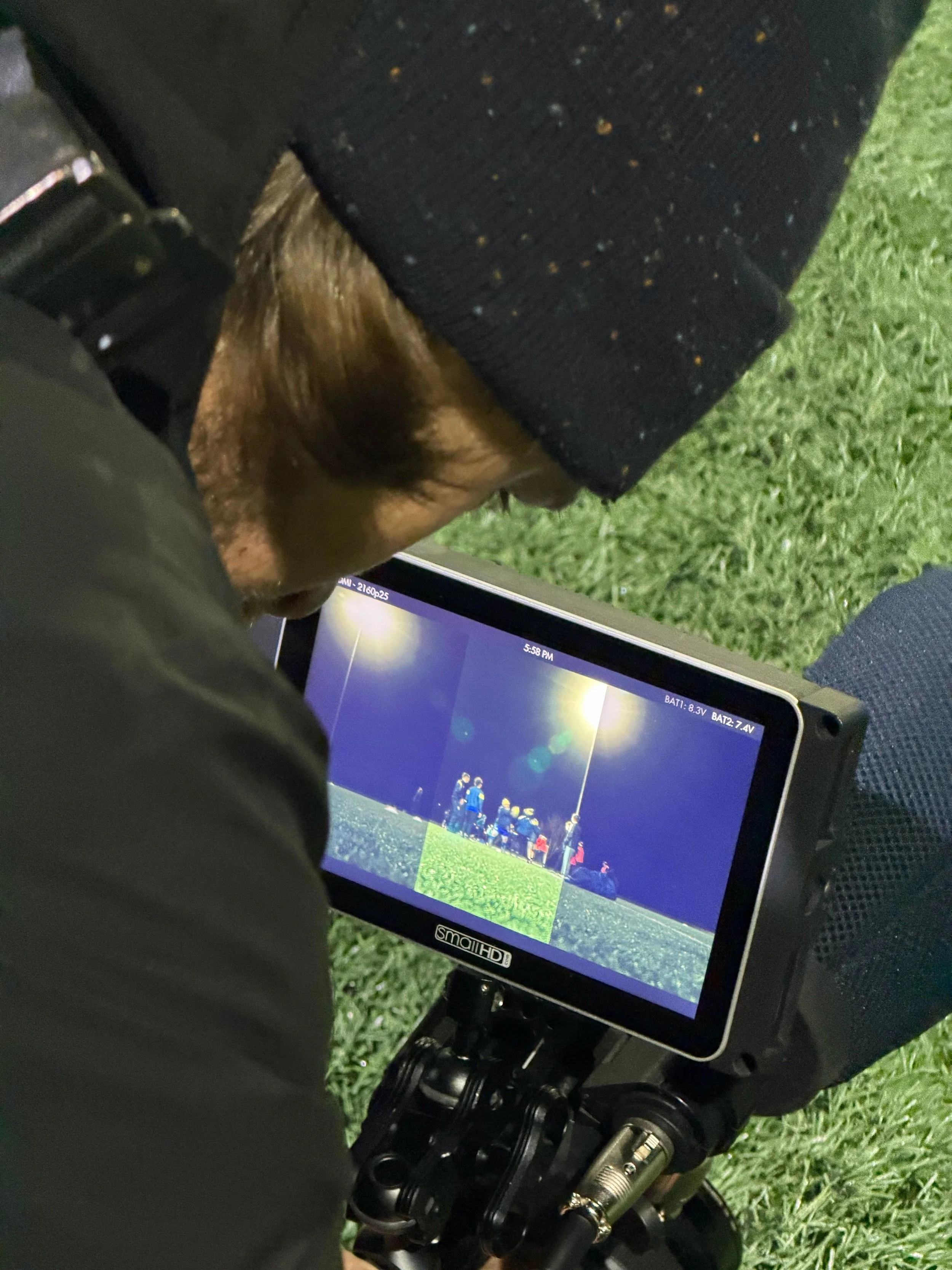 A person wearing a black beanie and black jacket, playing football at night under stadium lights, is viewing footage on a professional camera monitor on a tripod, with green grass in the background.