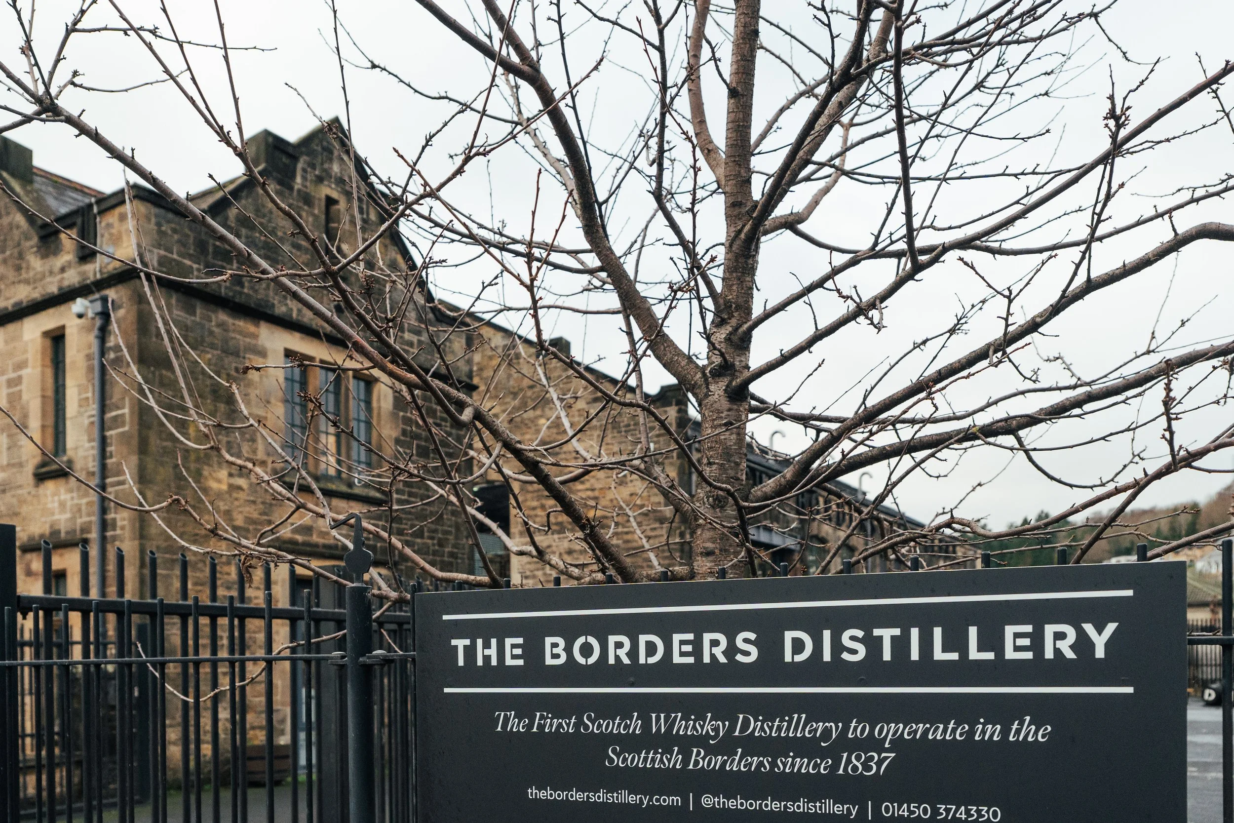 A sign for The Borders Distillery in front of a leafless tree with a stone building in the background.