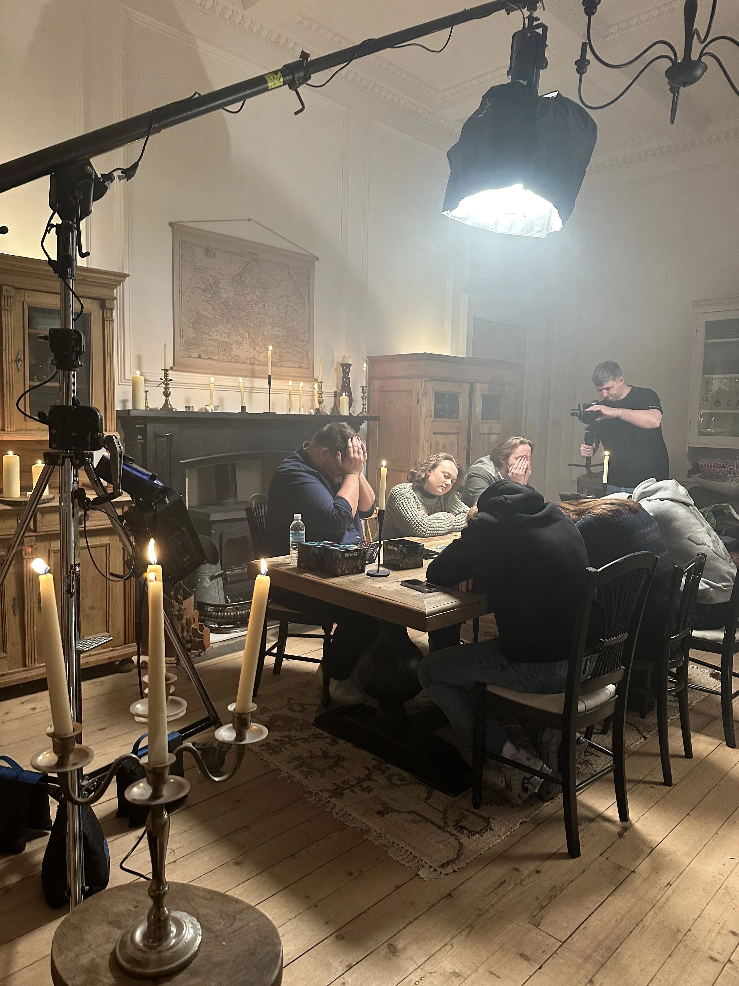 A group of people sit around a dining table during a filming scene, with candles on the table and a film crew member in the background adjusting equipment, in a room with wooden furniture and a map on the wall.
