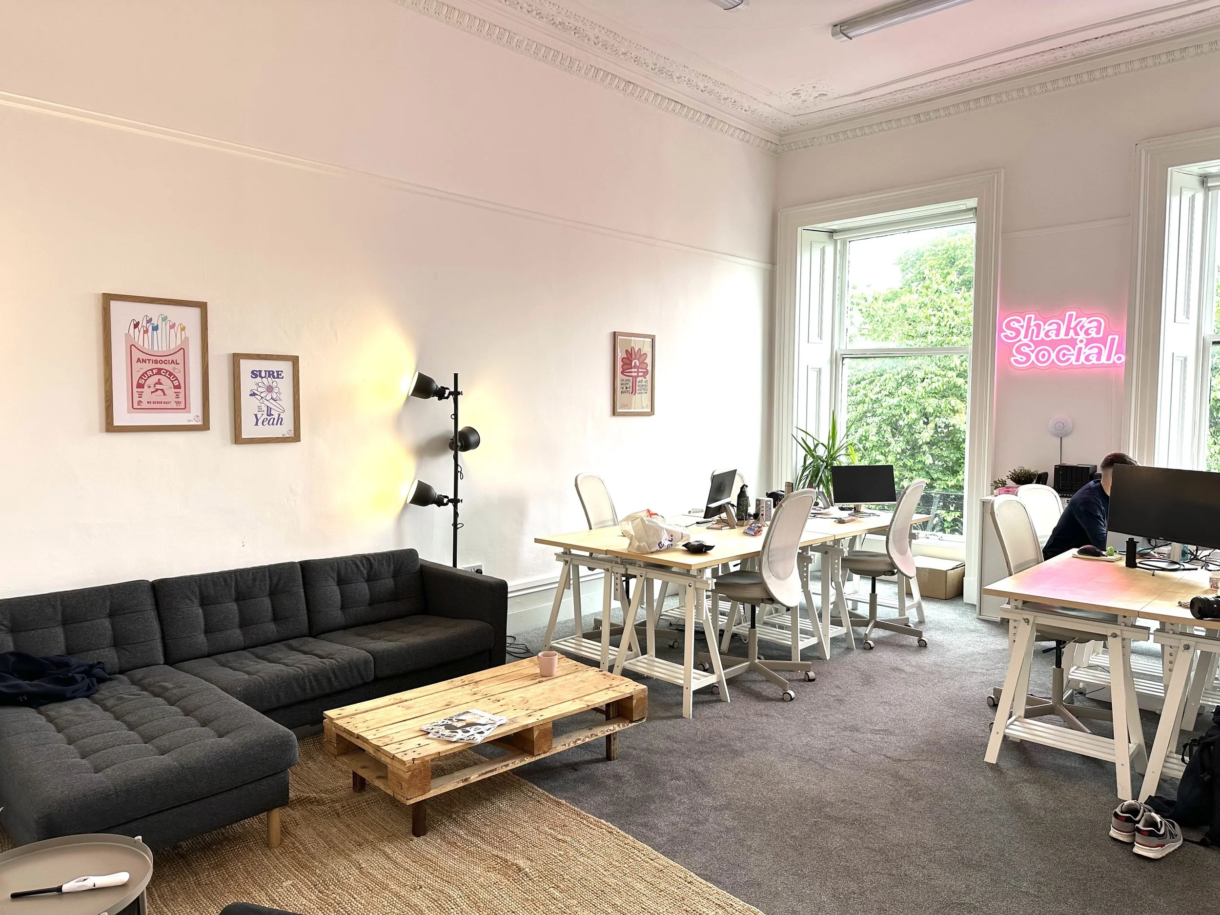 Open office space with white walls, a black sofa, a wooden pallet coffee table, and a floor lamp. Multiple desks with computers and chairs near large windows with greenery outside. A pink neon sign on the wall reads "Shaka Social."