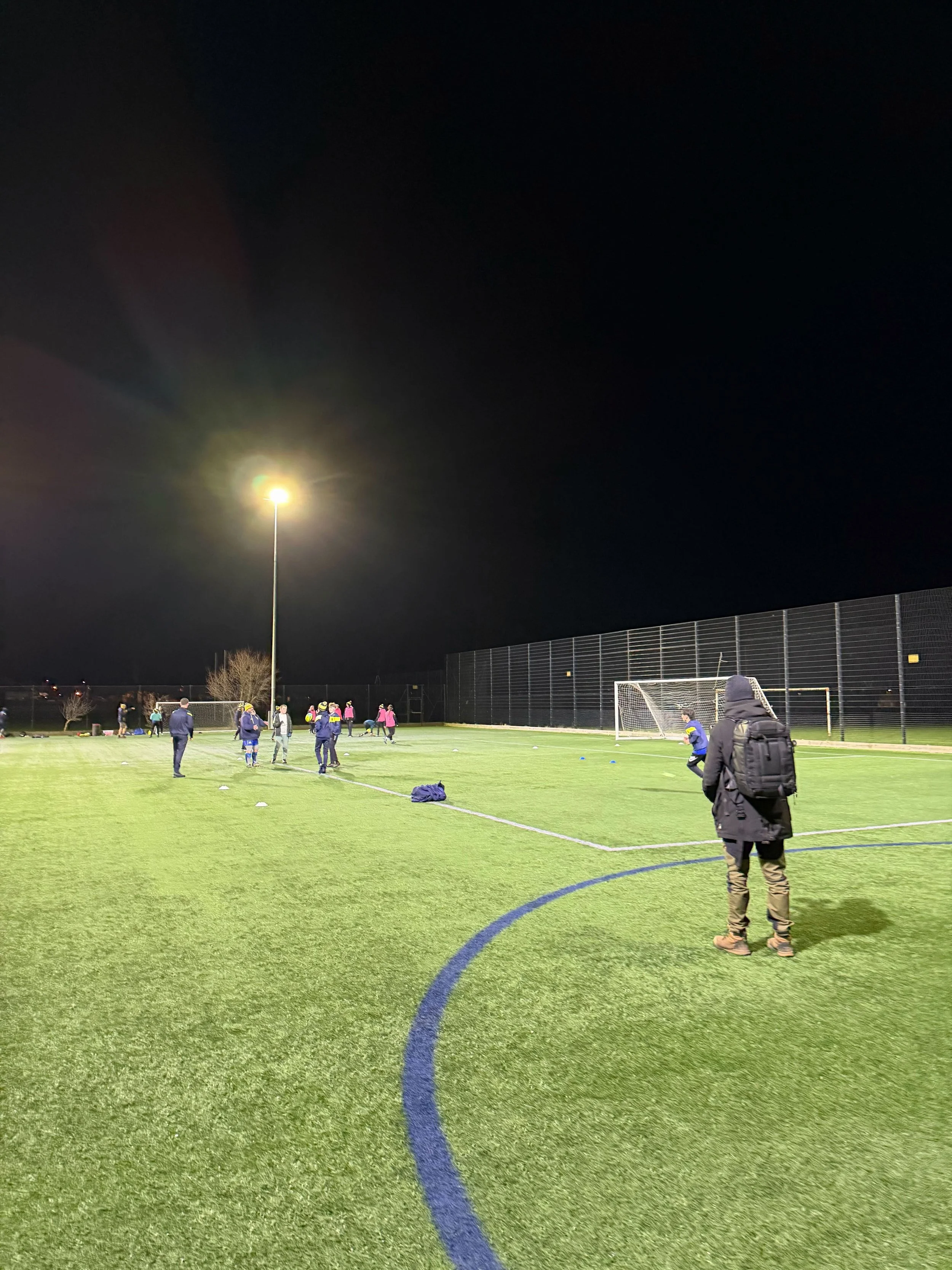 People practicing soccer on an outdoor field at night under bright overhead lights, with some individuals wearing sports attire and backpacks.