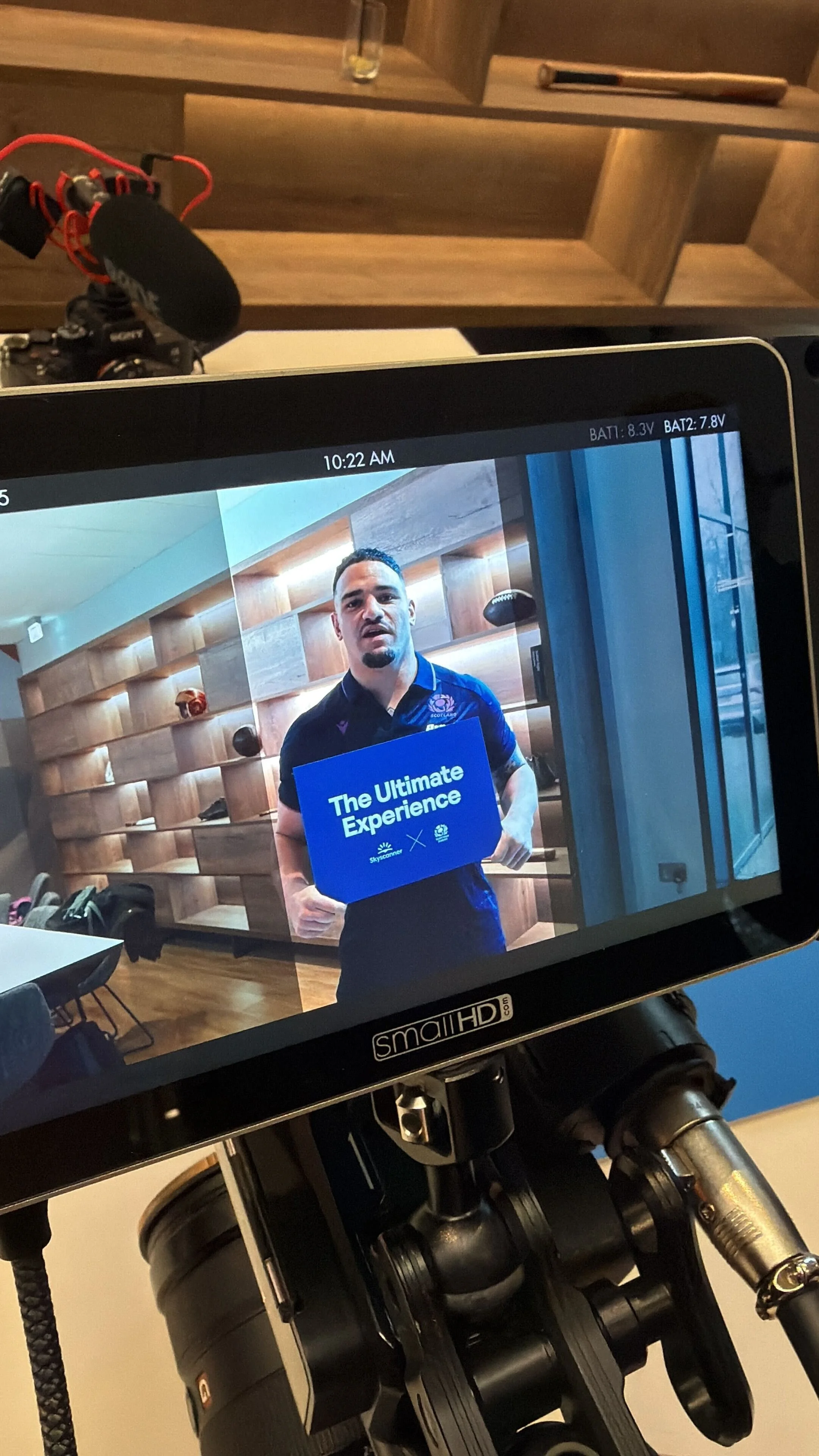 A man holding a sign that says "The Ultimate Experience" while being recorded on a camera monitor in an indoor setting with wooden walls and some tables and chairs.