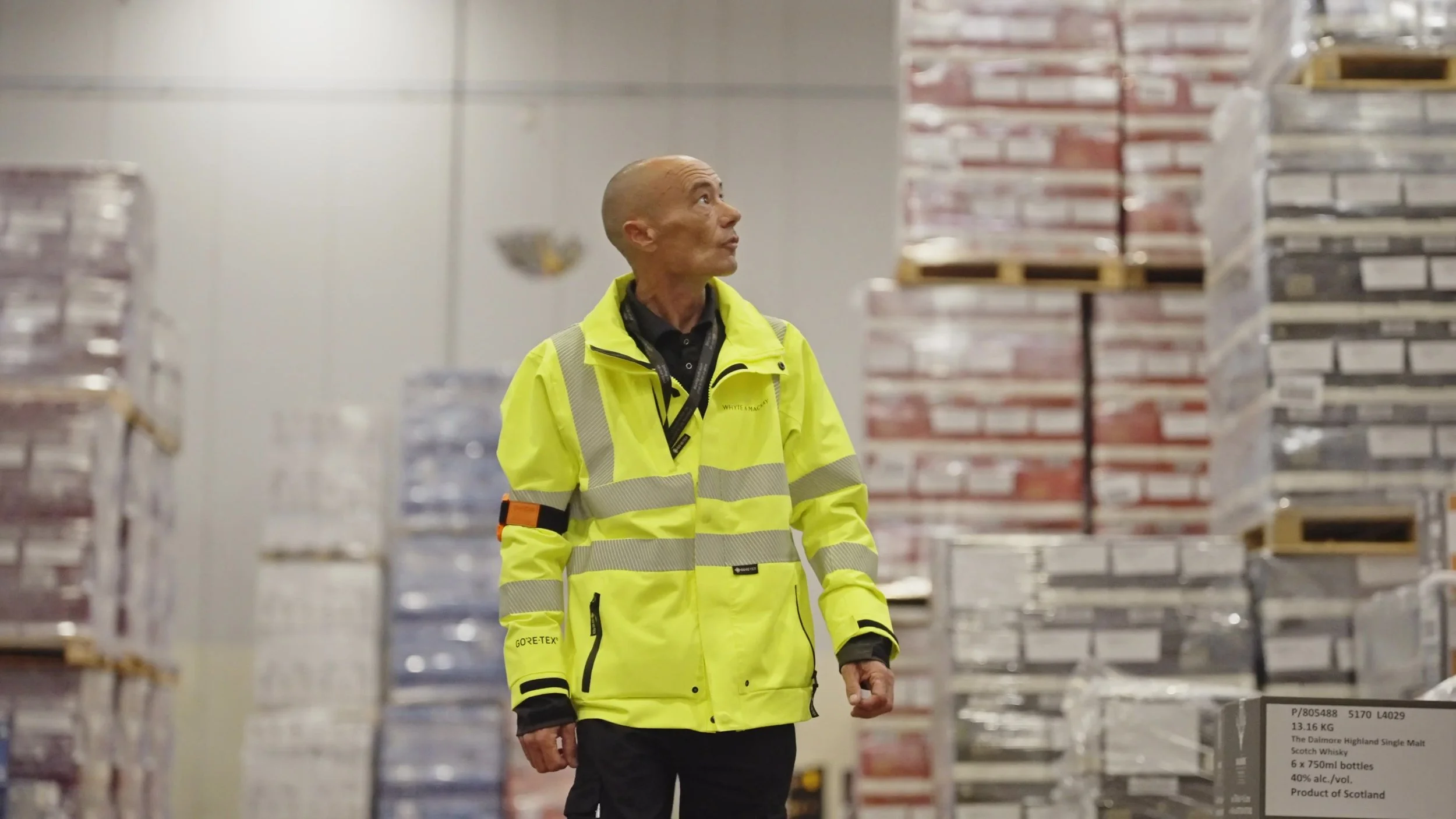 A man in a high-visibility yellow jacket looking around in a warehouse filled with stacked pallets of whisky bottles.