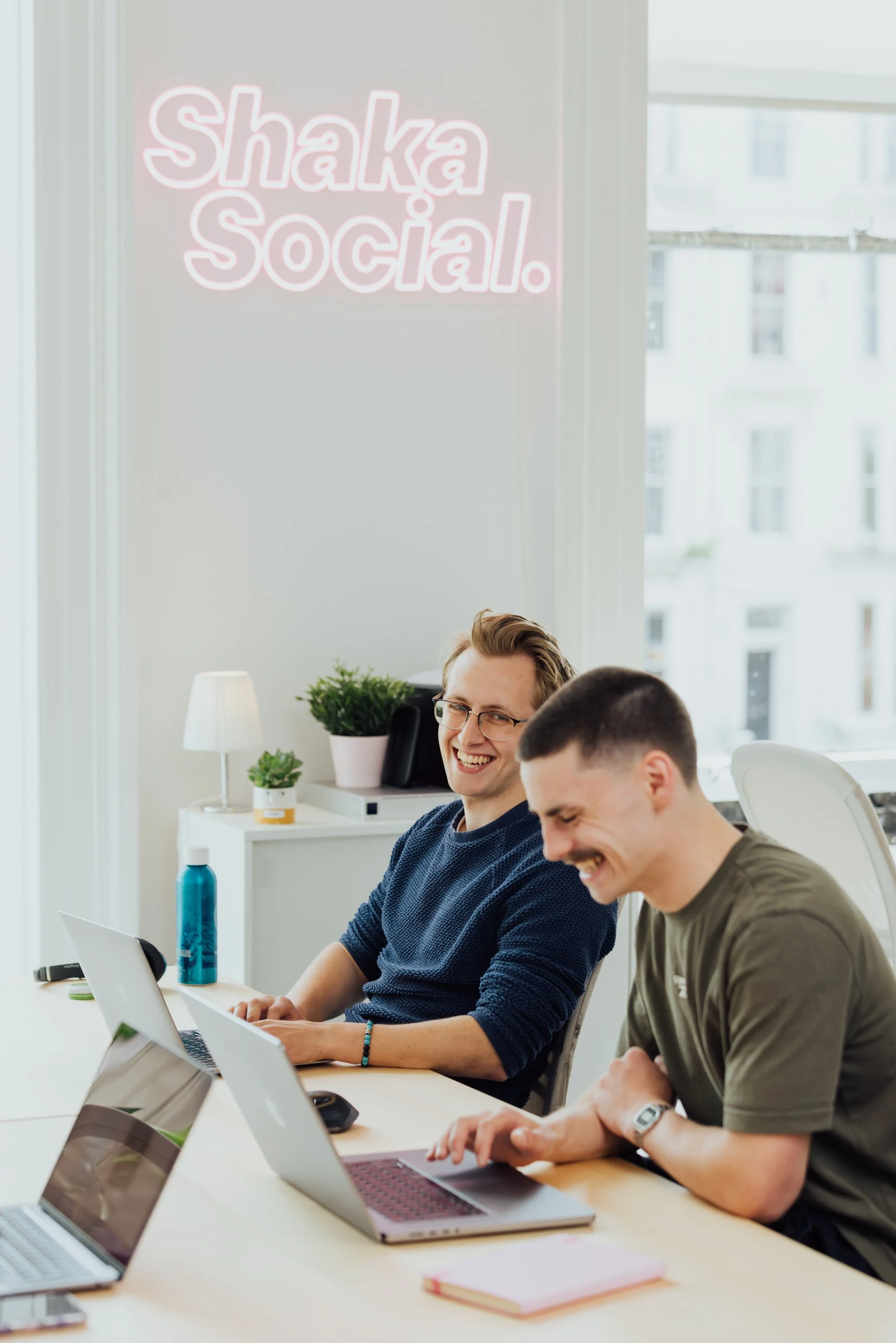 Two young men working at a desk, smiling, with laptops and notebooks in a bright office. A neon sign on the wall reads 'Shaka Social.'