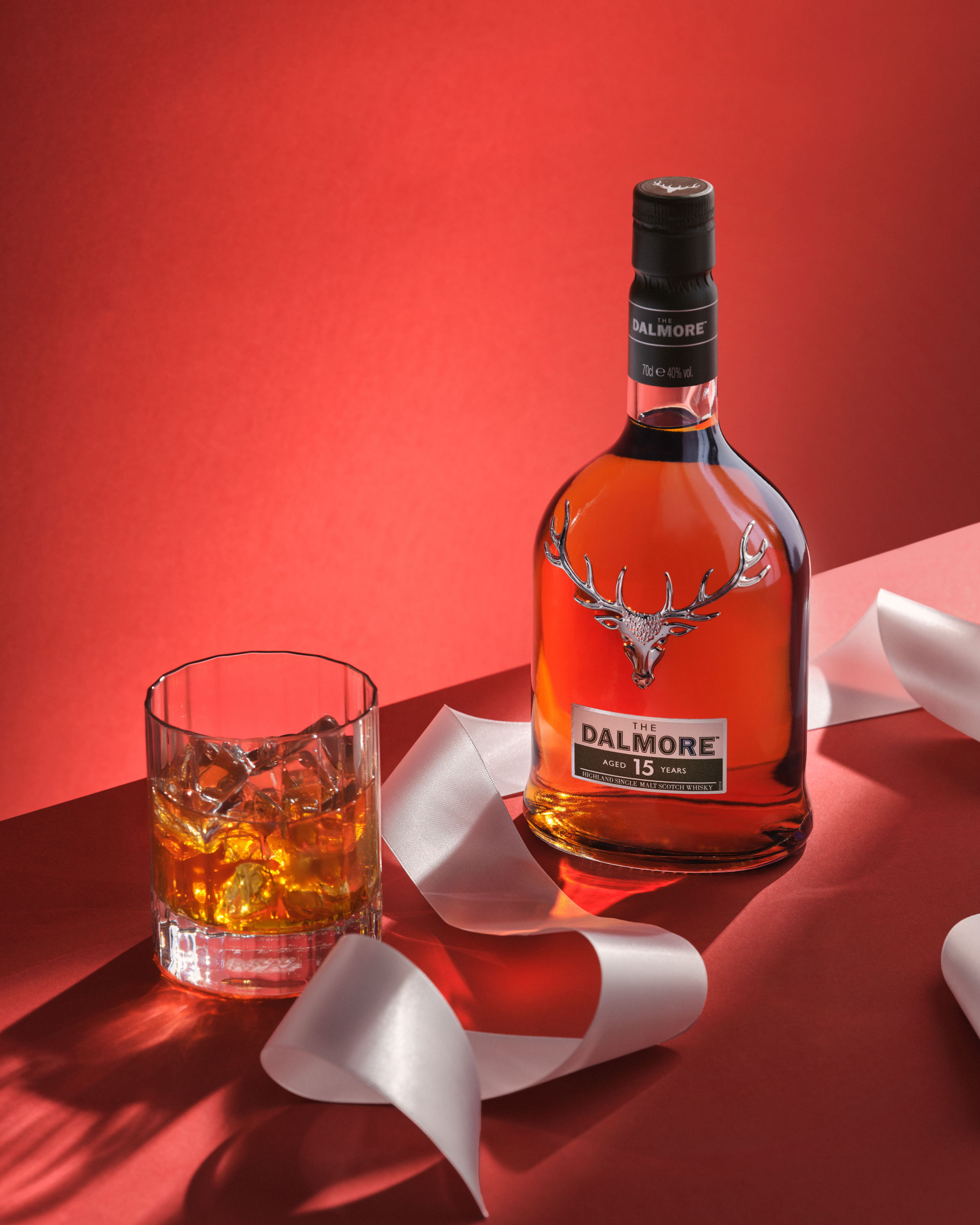 A bottle of The Dalmore 15-year-old Scotch whisky with a glass of whisky and ice cubes on a red surface, decorated with white ribbon and a red background.