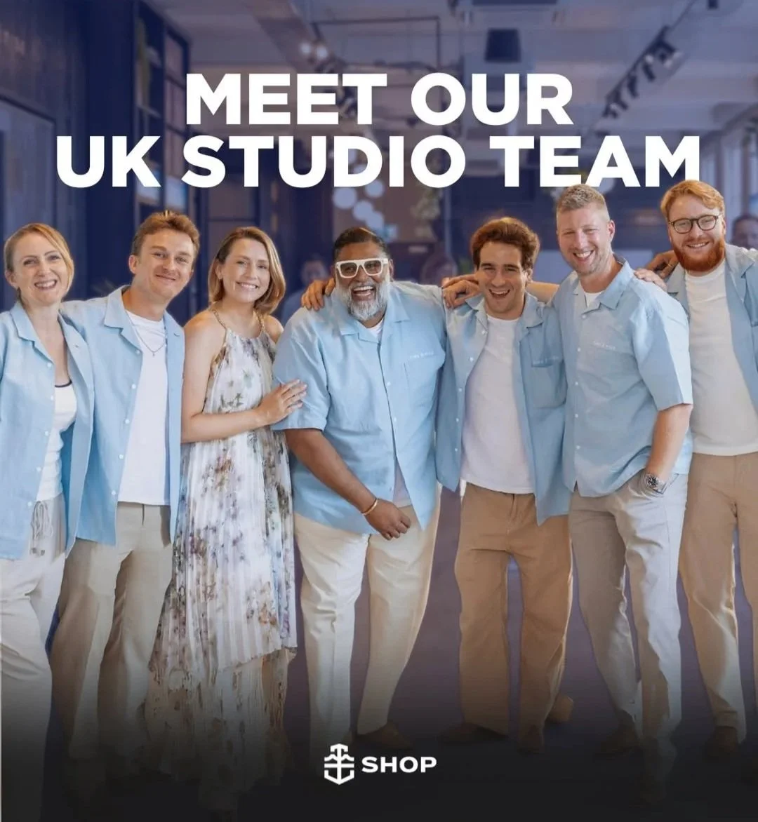 A diverse group of seven cheerful people wearing light blue shirts and casual pants standing together with arms around each other in an office or studio setting, smiling at the camera. The text overlay reads "Meet Our UK Studio Team," with a shop logo at the bottom.