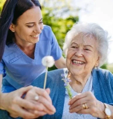 A young woman and an elderly woman smiling outdoors, holding flowers, with the younger woman assisting the elder.