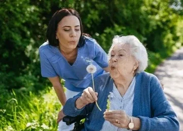 A young woman and elderly woman outdoors, with the elderly blowing on a dandelion.