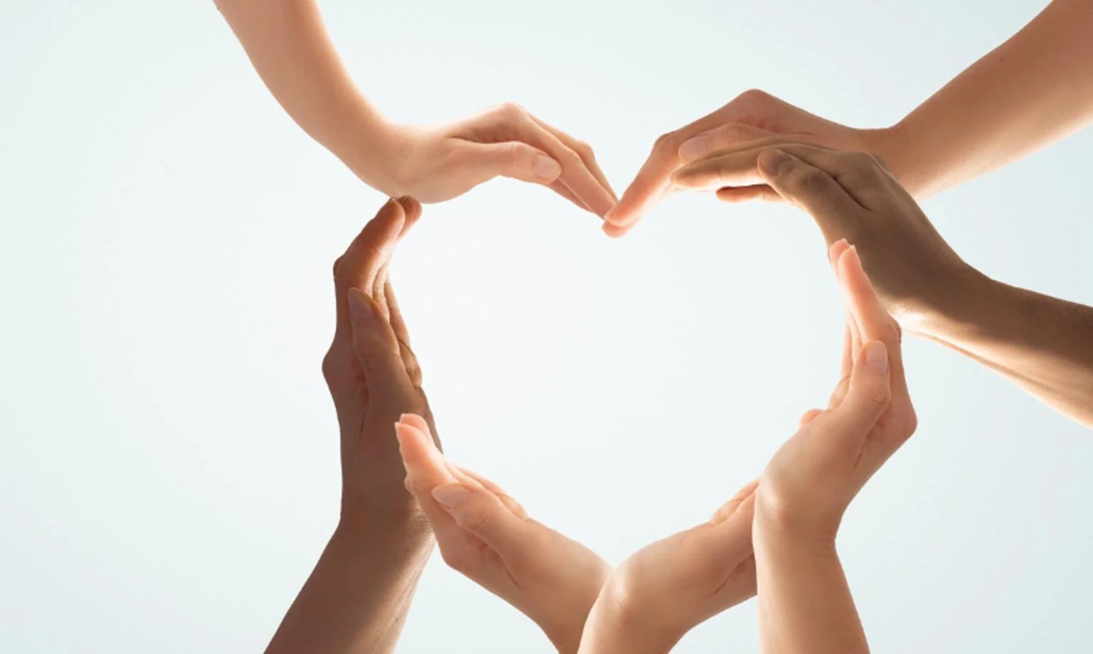 Multiple hands of diverse skin tones forming a heart shape against a light background.