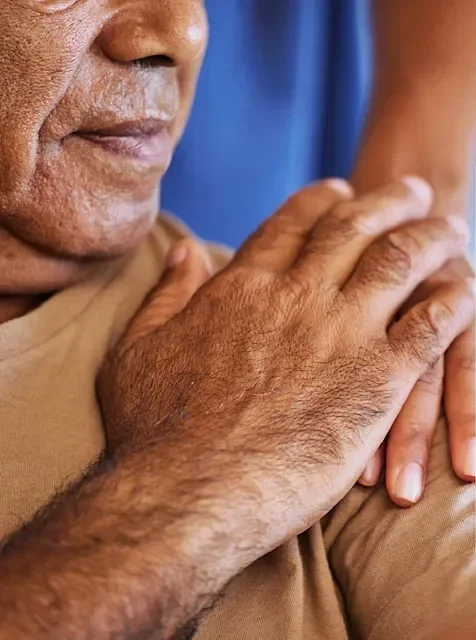Close-up of an elderly person's face and hand, with one hand resting on their shoulder, wearing a beige shirt.
