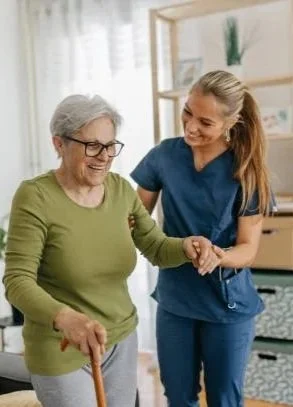 A nurse assisting an elderly woman with a cane, smiling in a caregiving setting.