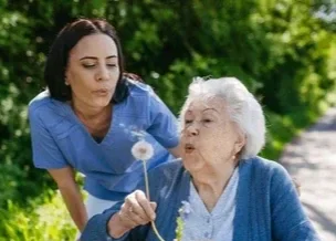 A young woman in blue scrubs standing beside an elderly woman blowing dandelion seeds outdoors with greenery in the background.