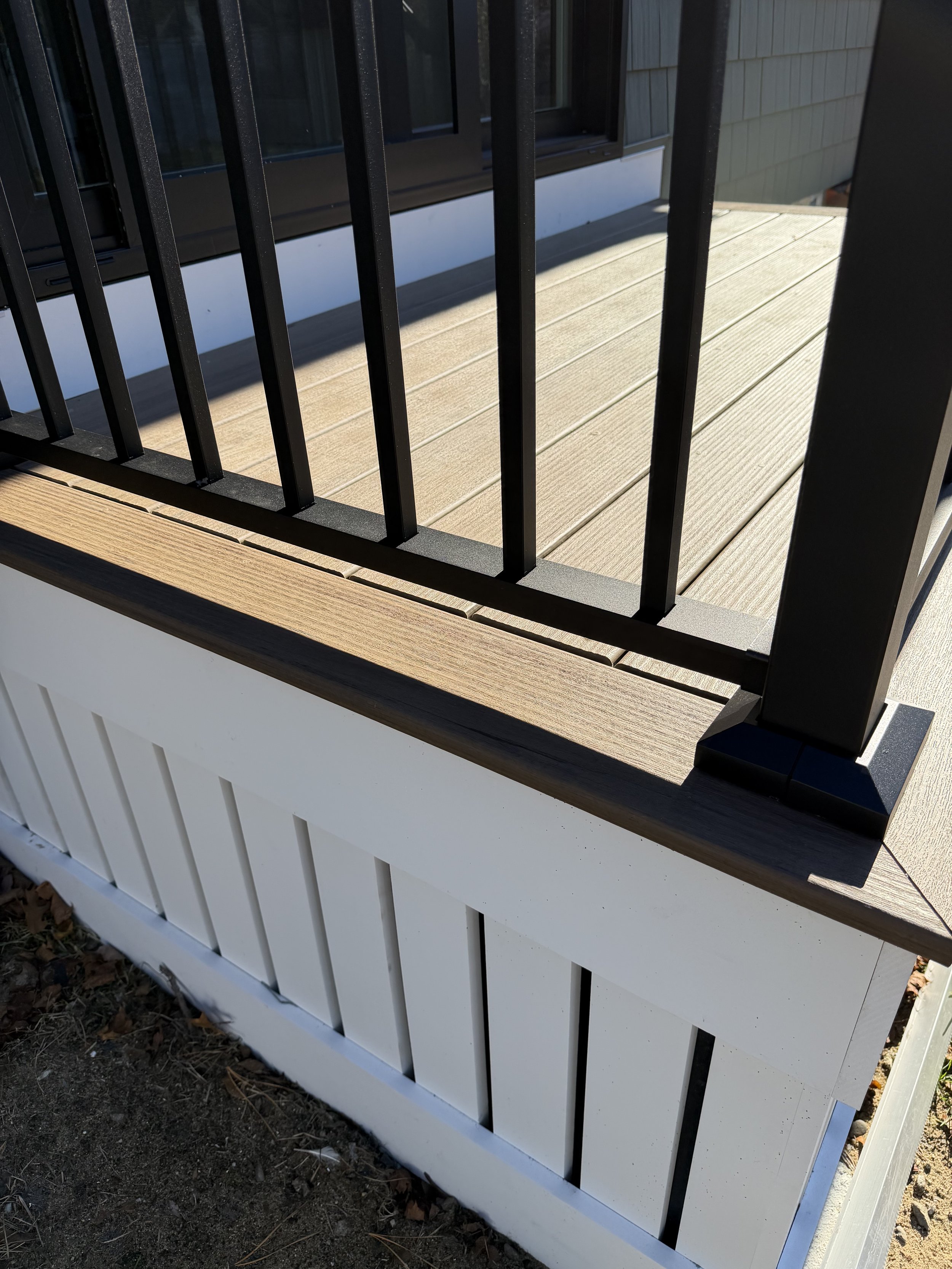 Close-up view of a wooden porch railing with black metal balusters, mounted on a white skirting with vertical slats, outside a building with dark gray siding.