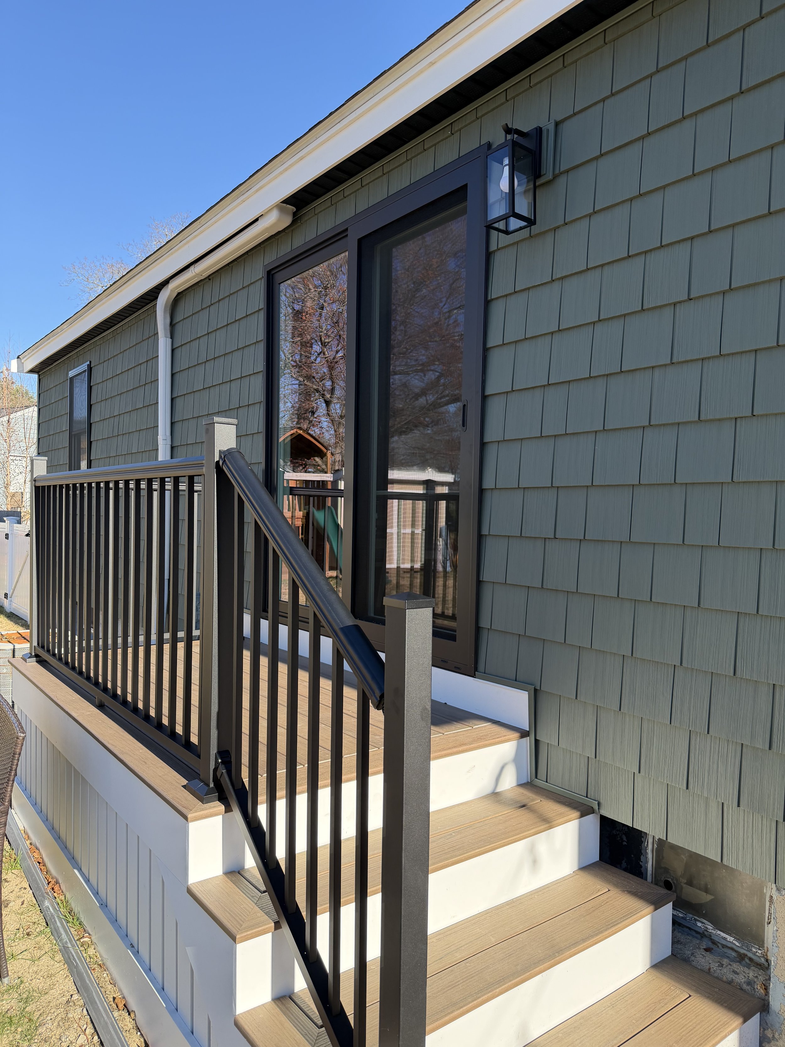 A small outdoor wooden deck with black metal railing, accessed by a three-step staircase, attached to a house with green siding and large sliding glass door. A wall-mounted outdoor lantern light is next to the door.