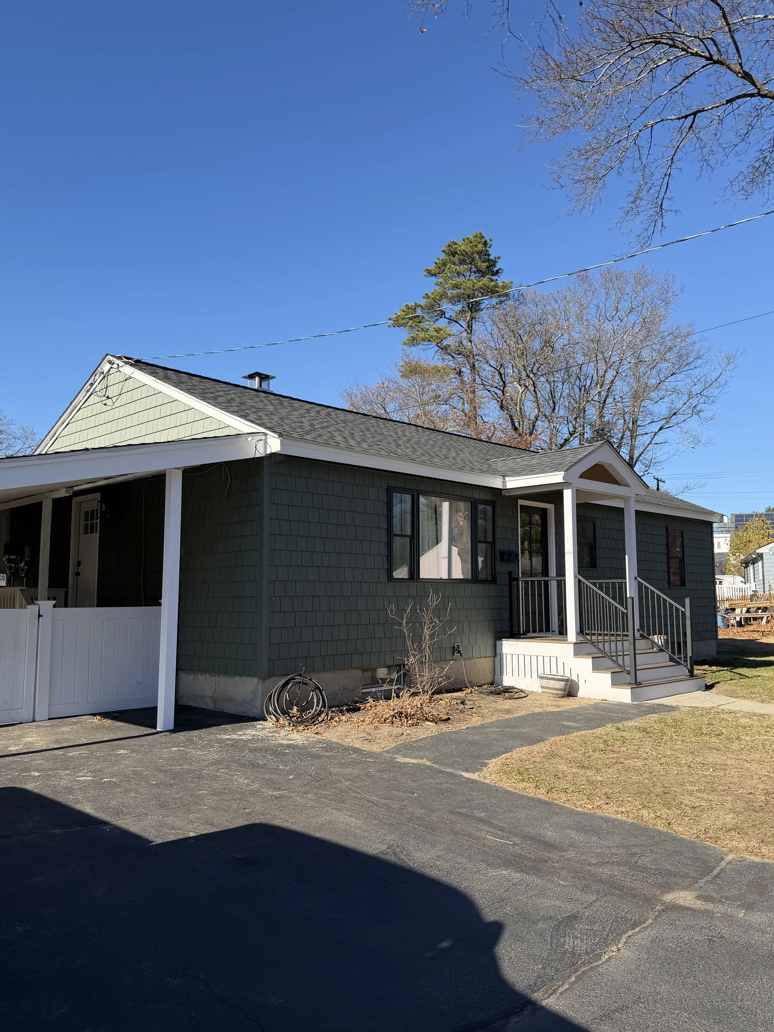A green house with a dark roof and small front porch with stairs, located on a paved driveway, with trees and a blue sky in the background.
