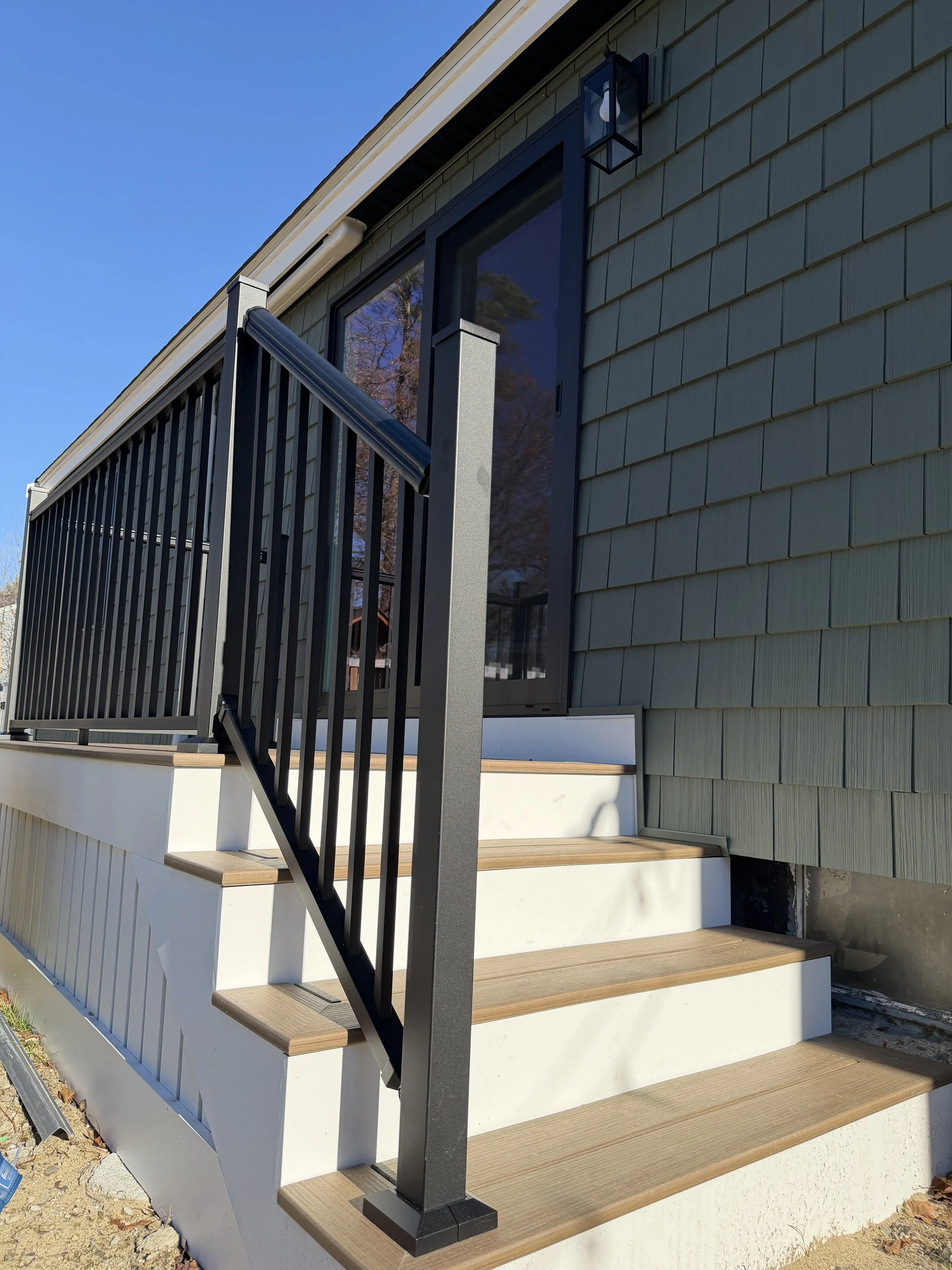 Exterior view of a house with new steps leading to a sliding glass door, featuring light wood tread stairs with black metal railing, green shingle siding, and an outdoor wall light fixture.
