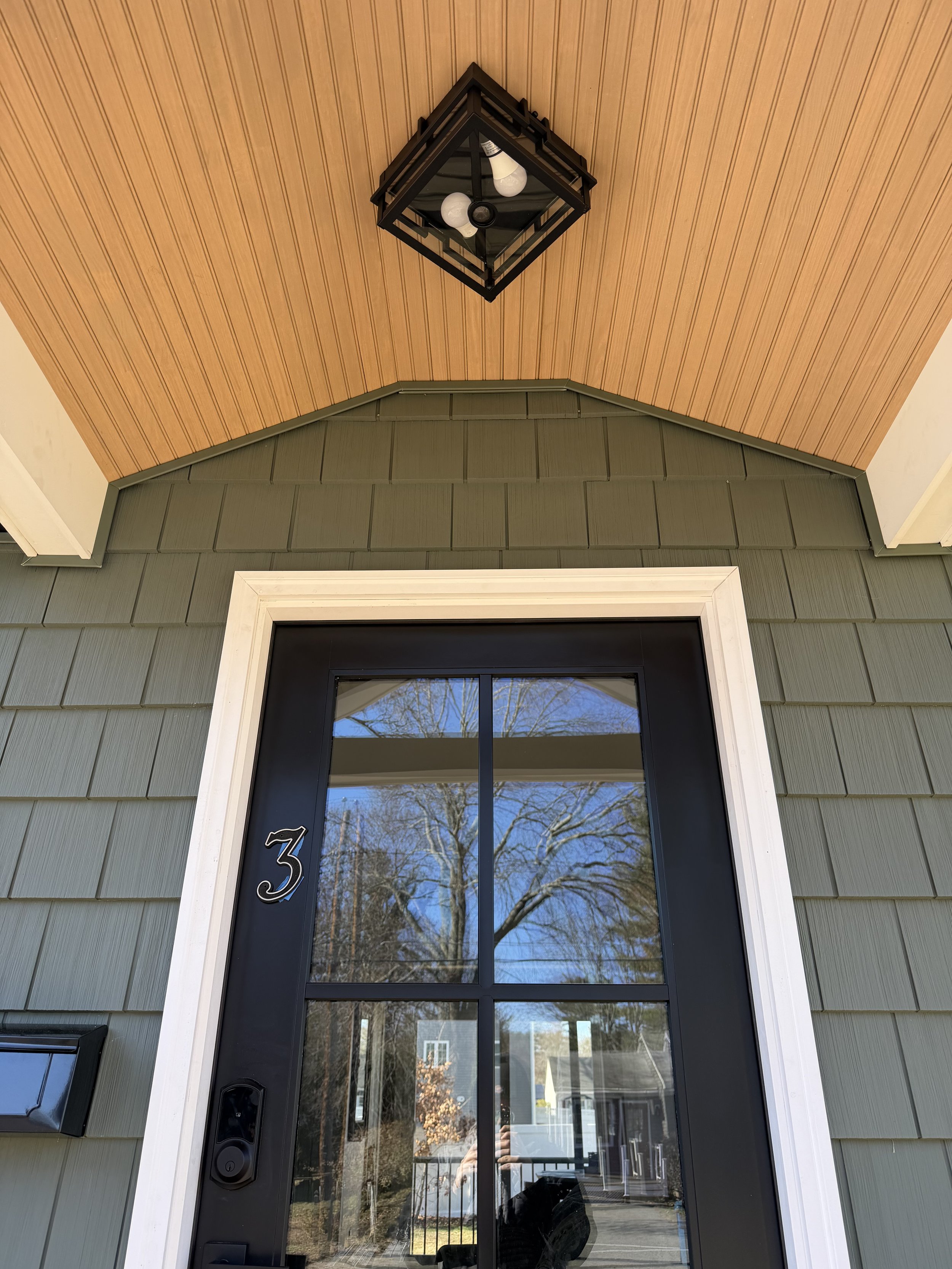 Front door with a glass panel reflecting trees and houses, framed in white trim, with a house number 3 on the door and a black doorbell to the left. The house exterior has olive green siding and there is a black mailbox on the porch. Above the door, 
