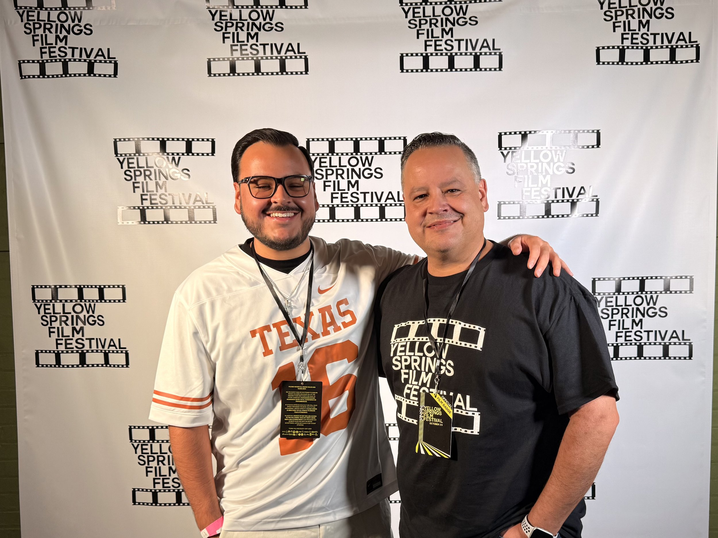 Two men standing in front of a backdrop with the words 'Yellow Springs Film Festival' and filmstrip patterns. They are smiling, with one man wearing a white Texas football jersey and the other wearing a black T-shirt. Both have lanyards around their necks.
