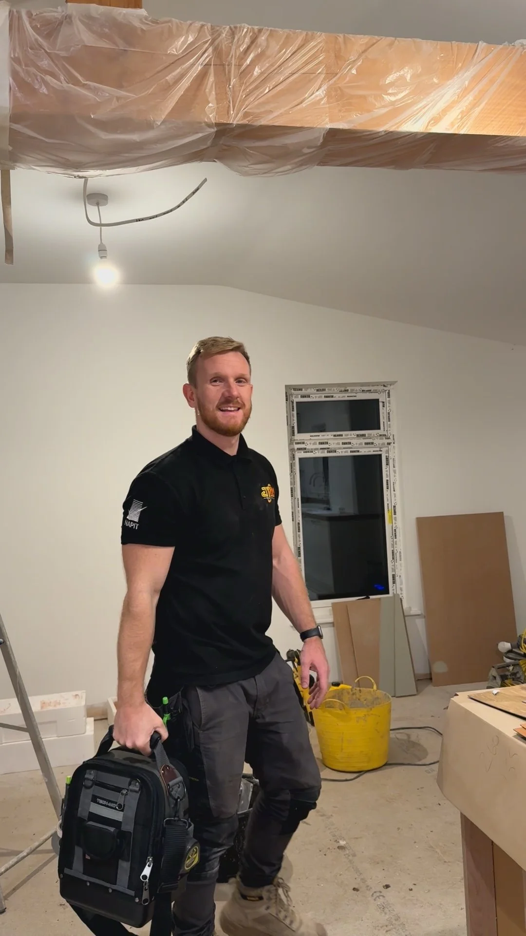 JTM Owner, Joe seen inside a room under construction, smiling, holding a black tool bag, with construction tools and materials around him