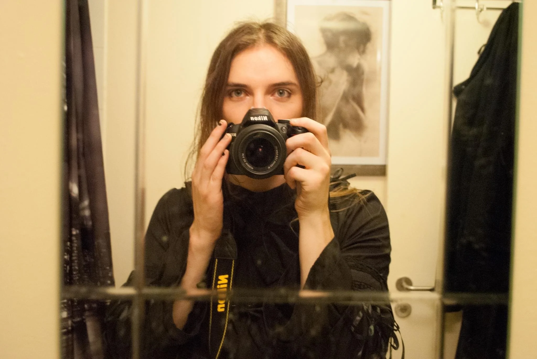 Drag artist taking a selfie in a mirror with a DSLR camera, brown hair, black clothing, in a room with a poster of a woman in the background.