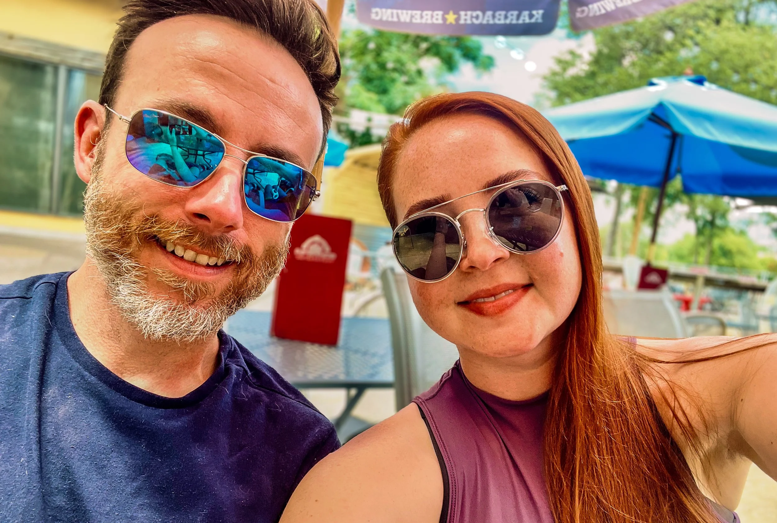 A man and woman taking a selfie at an outdoor cafe, both wearing sunglasses and smiling. The background shows tables, chairs, umbrellas, and trees.