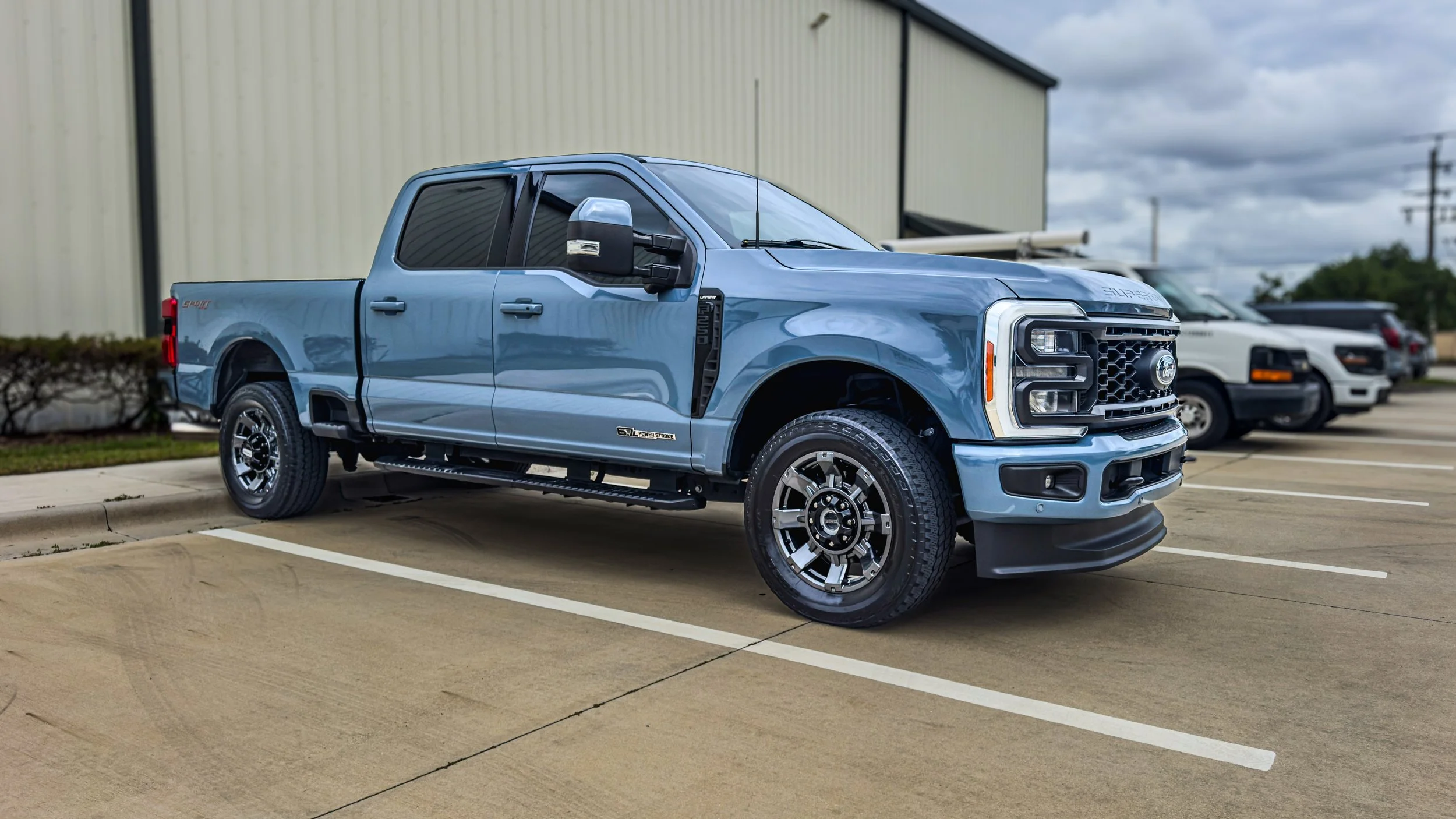 A light blue Ford pickup truck parked in a parking lot next to a beige industrial building, with other vehicles in the background under cloudy skies.