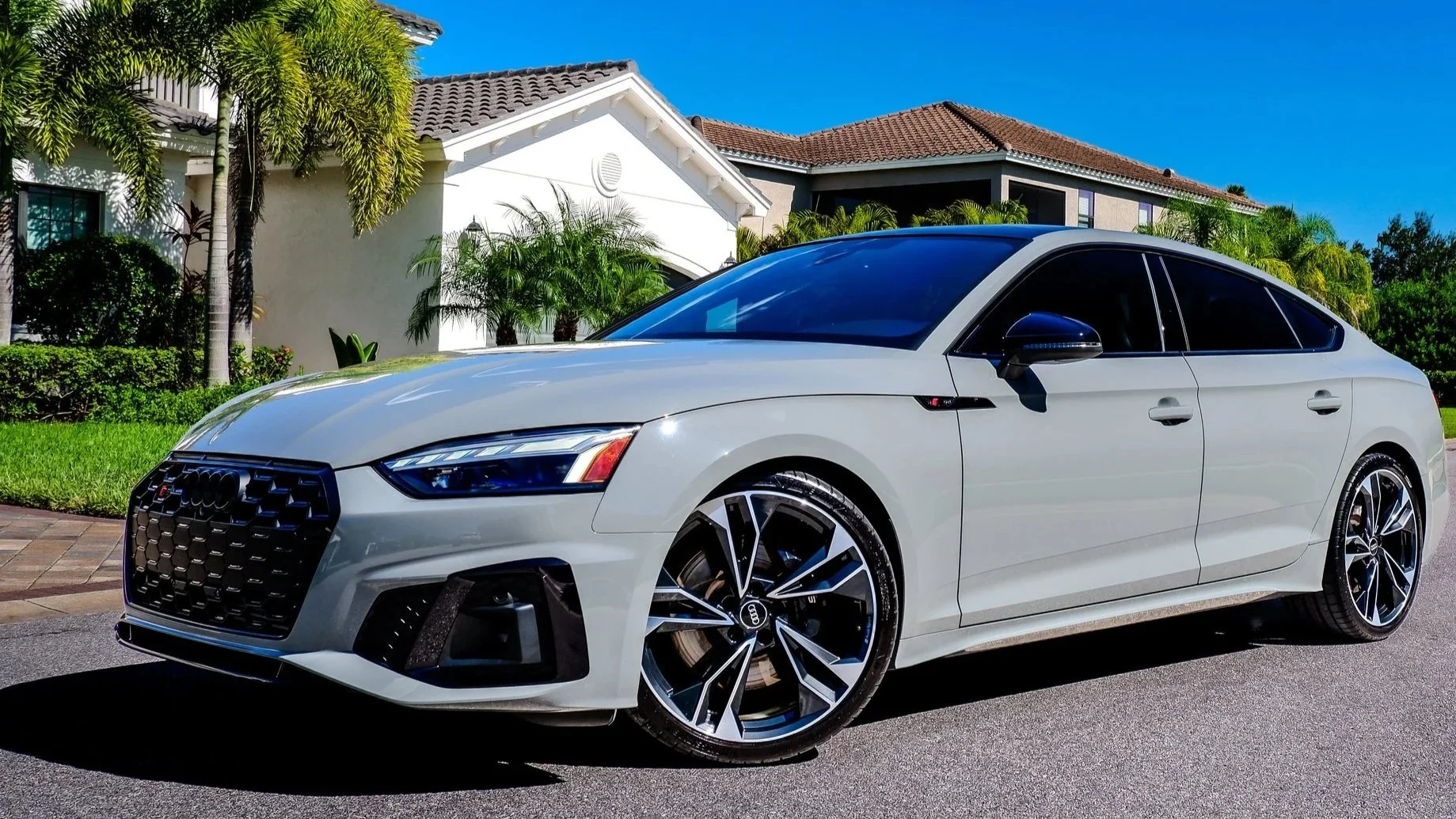 A white Audi sedan parked on the street in front of residential houses with lush greenery and palm trees, under a clear blue sky.