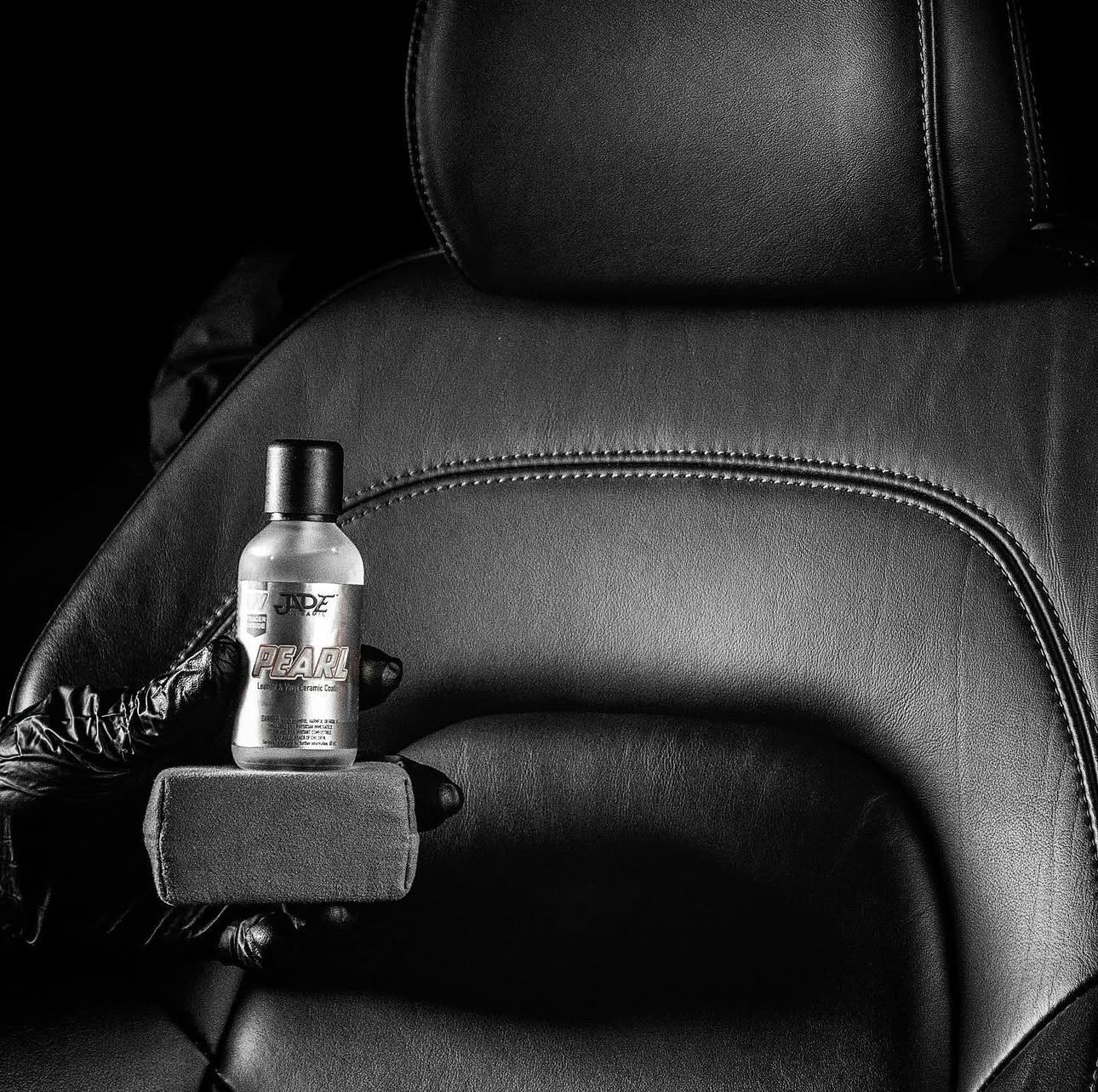A hand wearing a black glove holding a bottle of Jade Pearl ceramic coating over a black leather car seat with an armrest.