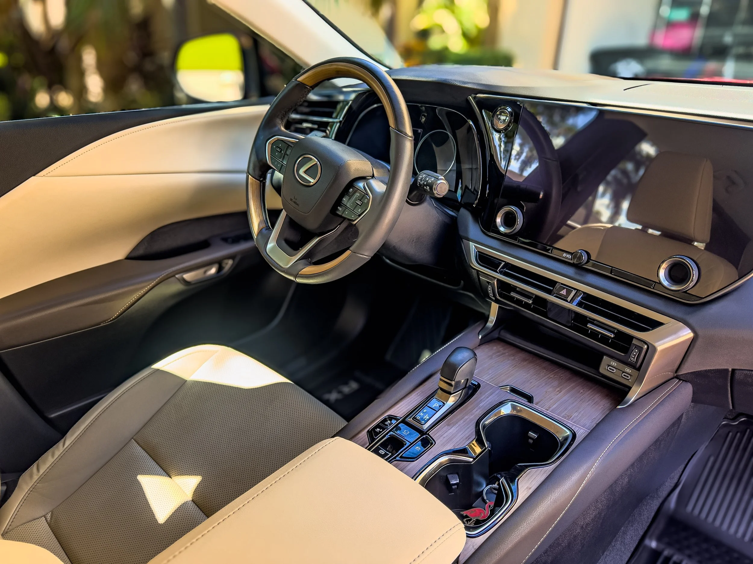 Interior of a luxury car, showing a steering wheel with a Lexus logo, a large digital display, dual air vents, a wooden center console with gear shift, and beige leather seats.