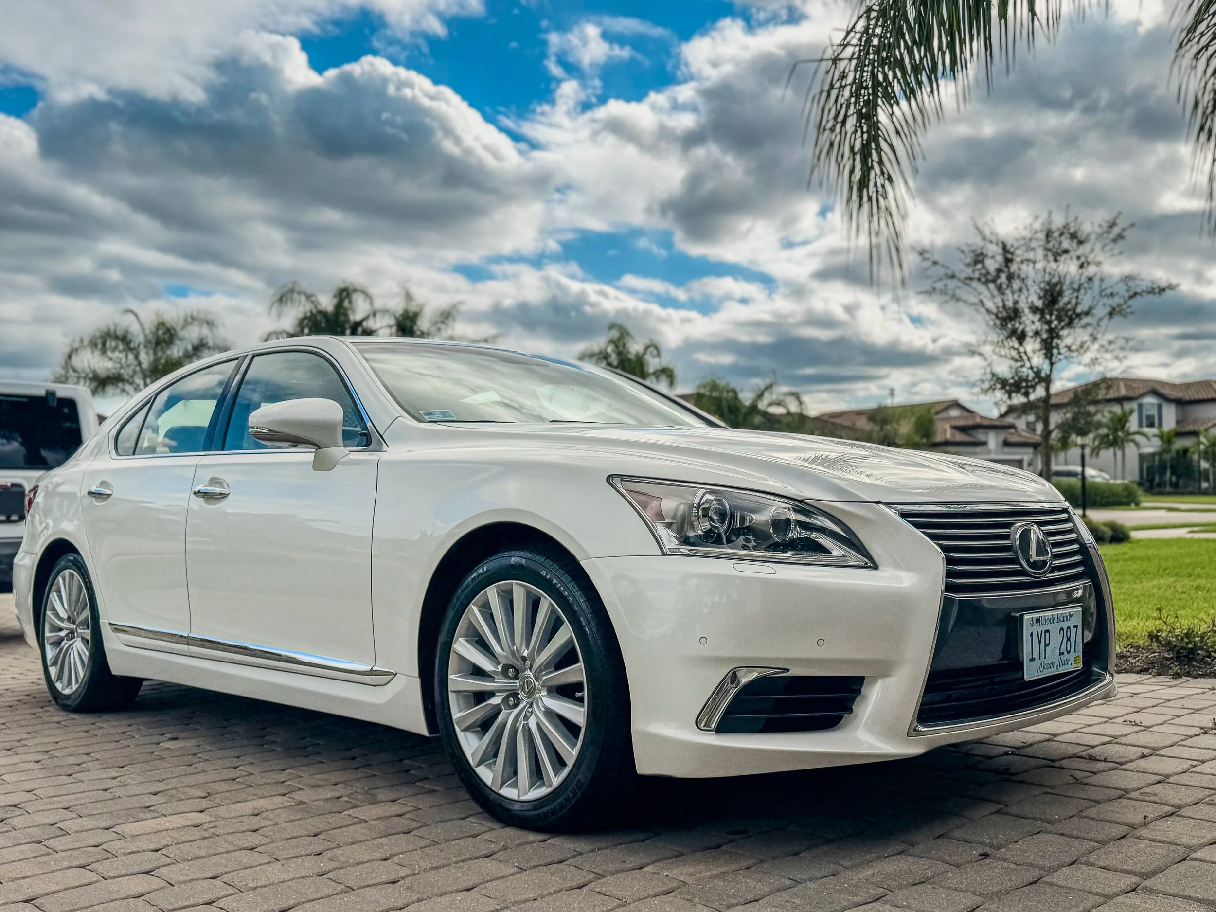 White Lexus sedan parked on a brick driveway under a partly cloudy sky with palm trees and houses in the background.