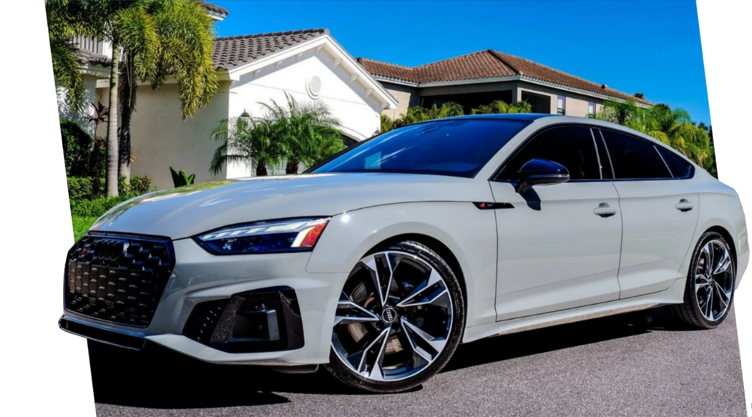 A white modern luxury sedan parked on a driveway in front of residential houses with palm trees under a clear blue sky.
