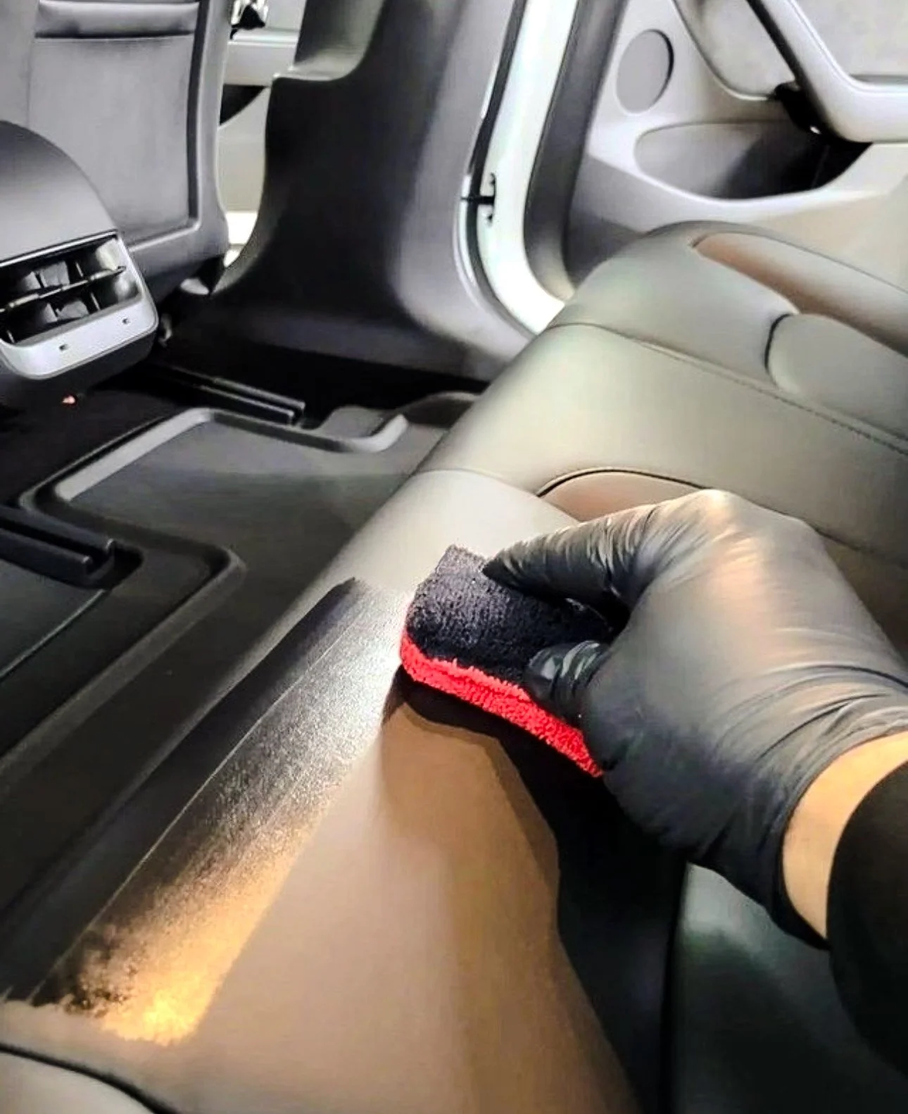 Person cleaning car interior's leather surface with a red and black cloth, wearing black gloves.