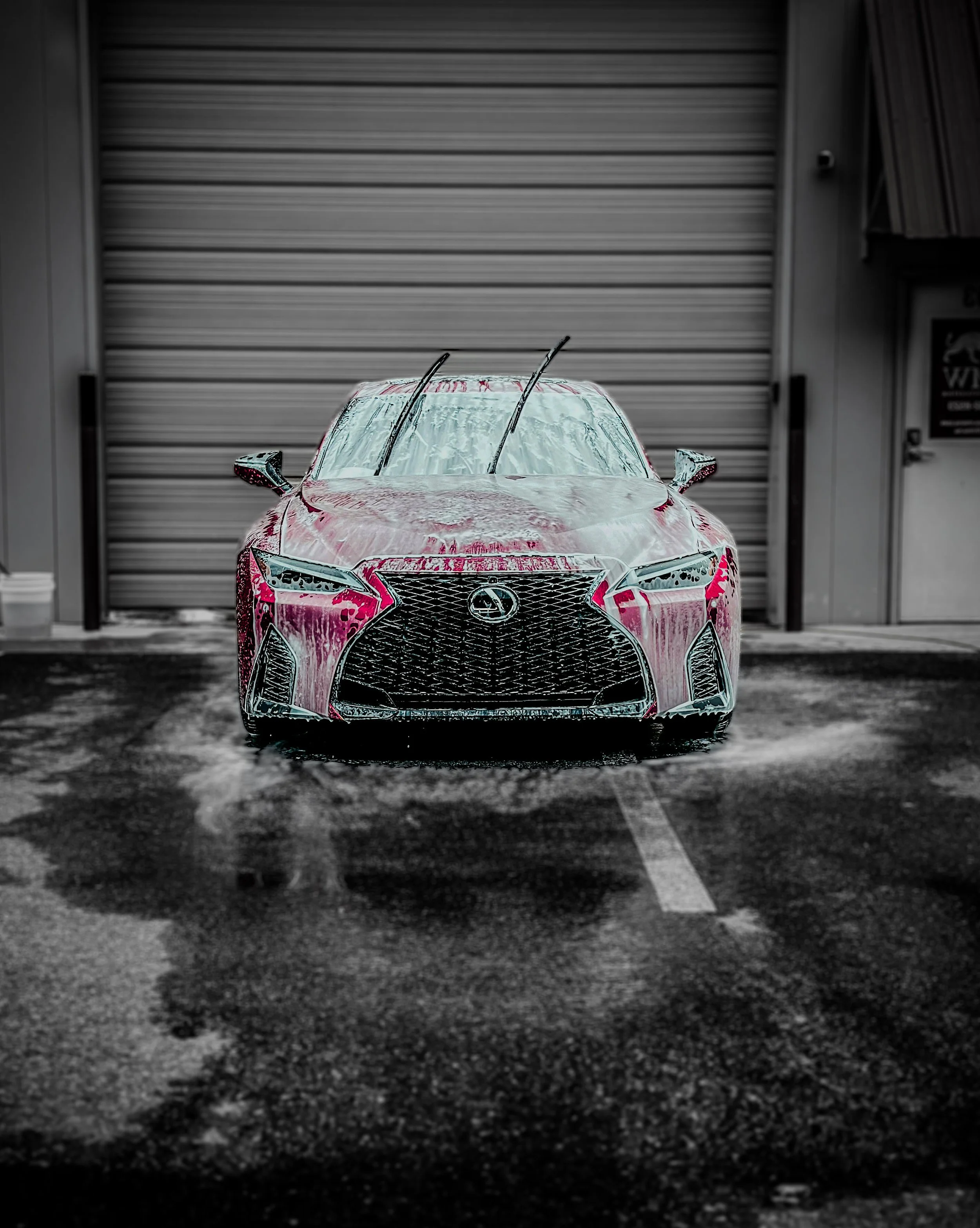 Red Lexus coupe parked in front of garage door with soap foam on the car, windshield wipers lifted, and wet pavement underneath.