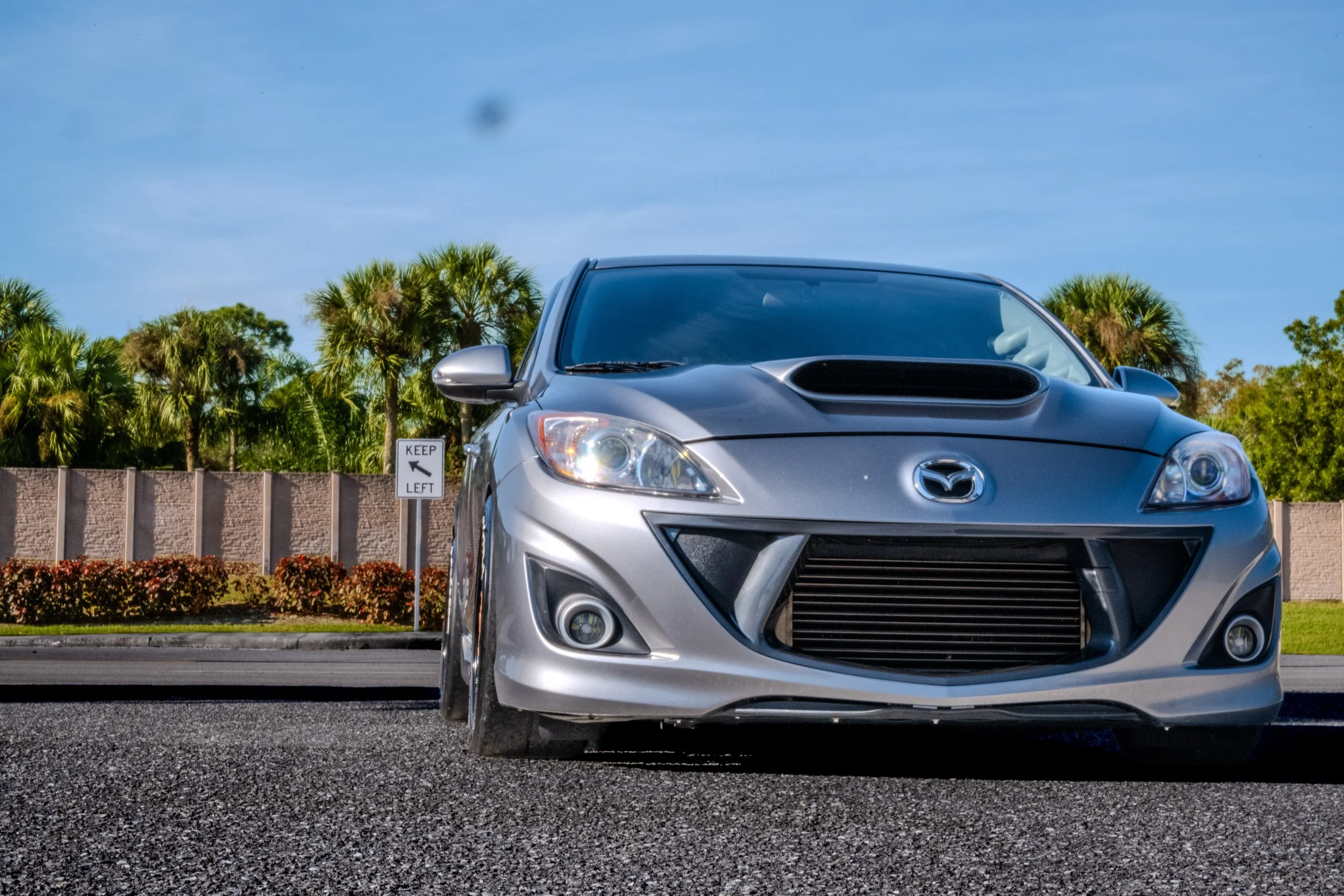 Front view of a silver Mazda car parked on the street with a palm-tree background and a 'Keep Left' sign on the roadside.