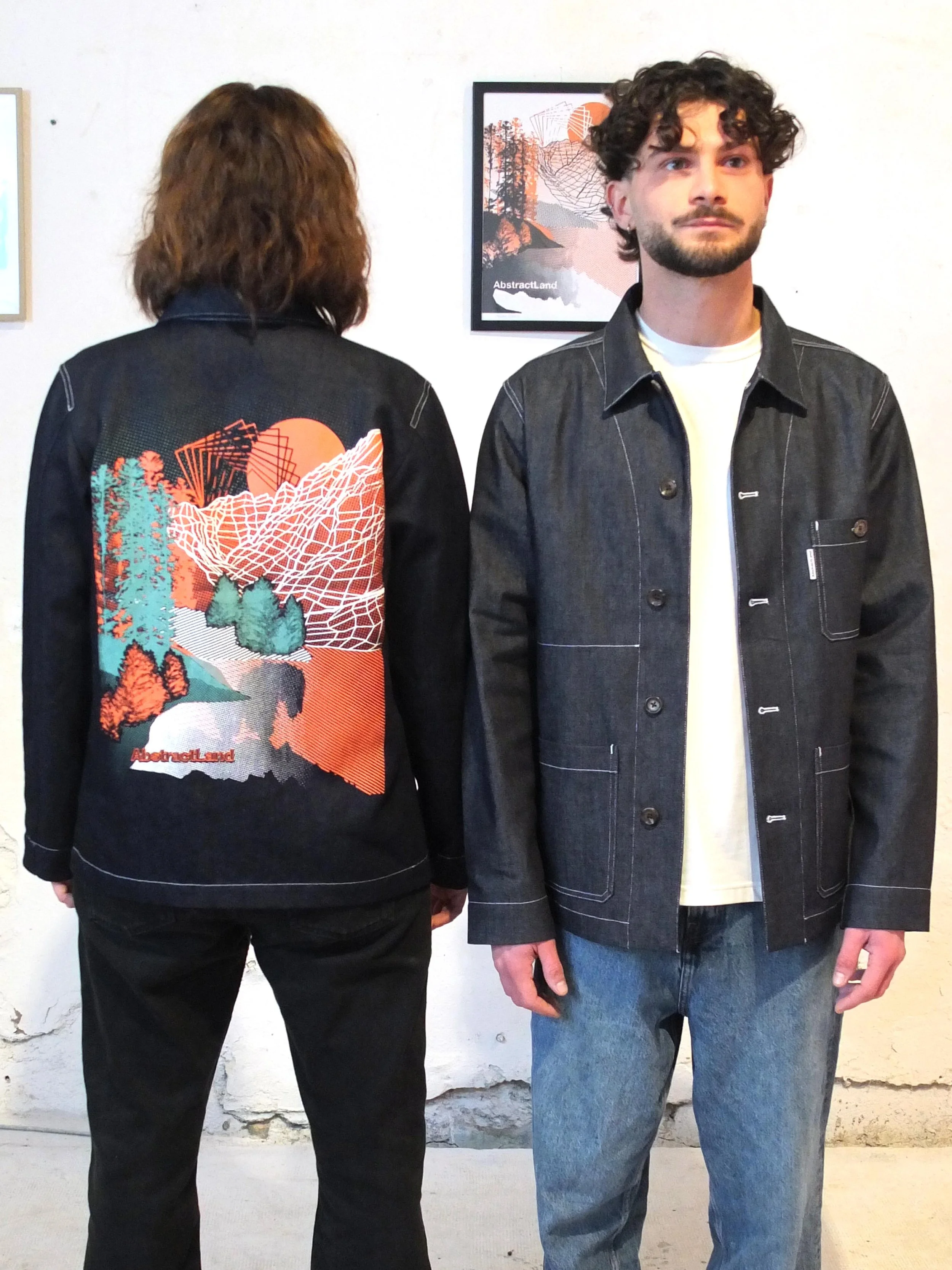 Deux personnes portant des vestes en denim dans une galerie d'art. La personne à l'avant a une veste noire avec un design graphique coloré dans le dos, et l'autre a une veste en denim simple. Une œuvre d'art est accrochée au mur derrière eux.