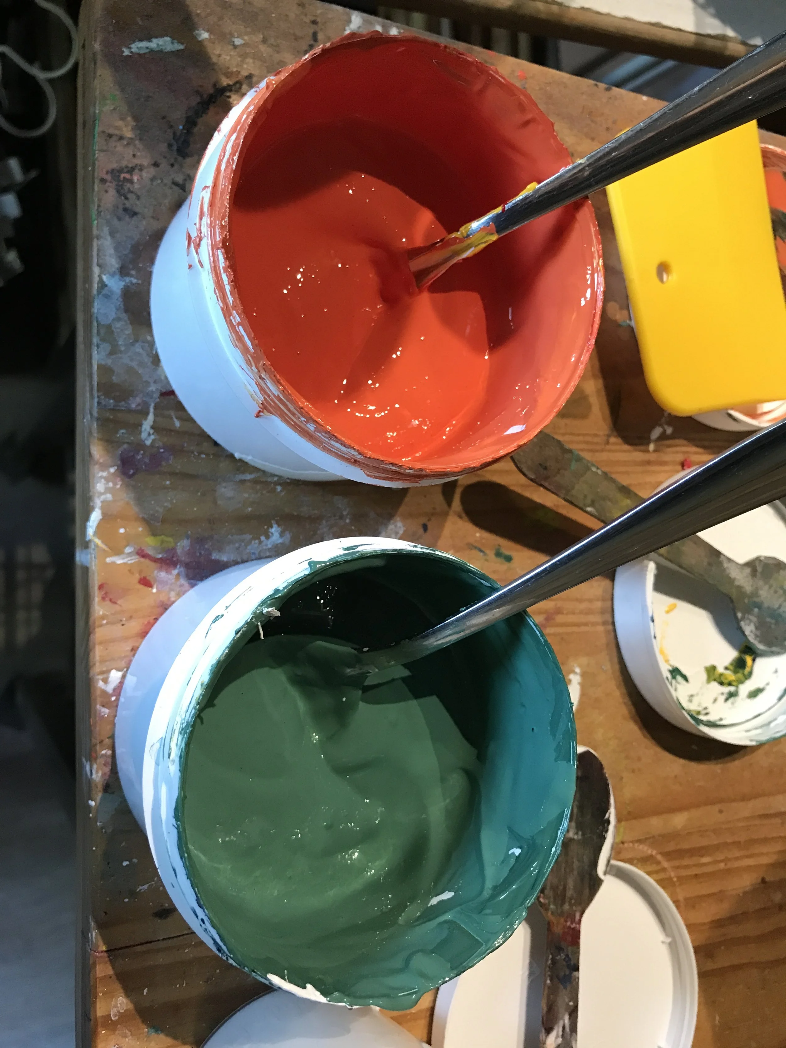 Deux pots de peinture, un rouge et un vert, avec des pinceaux dedans. La surface en bois est sale avec des taches de peinture.