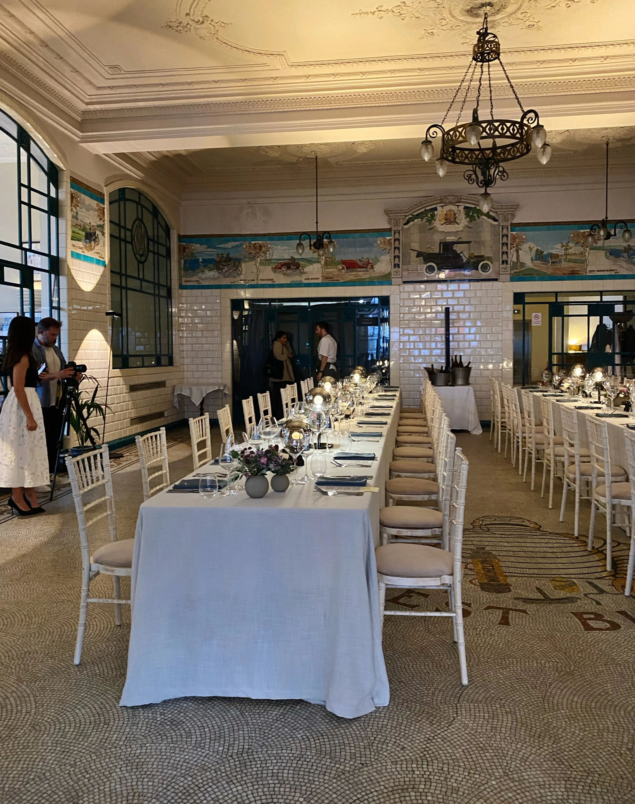 Elegant dining room with long table set for a formal event, decorated with glasses, plates, napkins, and floral centerpieces. Vintage chandeliers hang from the ornate ceiling, and large windows let in natural light. A mural with vintage cars decorate