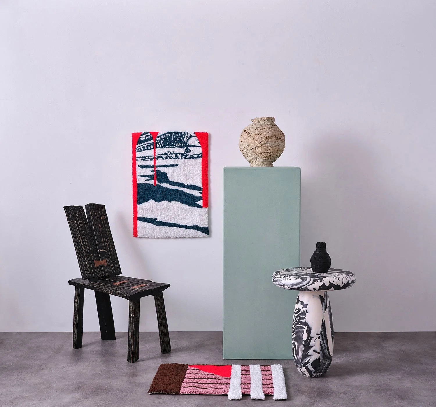 Interior art display with a black wooden chair, a wall hanging with blue, white, and red colors, a large pale blue pedestal with a textured beige vase, a black textured sculpture on a black and white marbled pedestal, and three small rugs on the floo