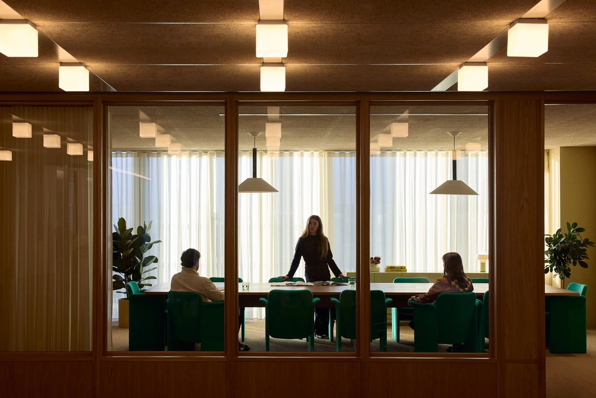 A woman standing at the head of a conference table in a modern meeting room, with two people seated on either side, and large windows with curtains in the background.