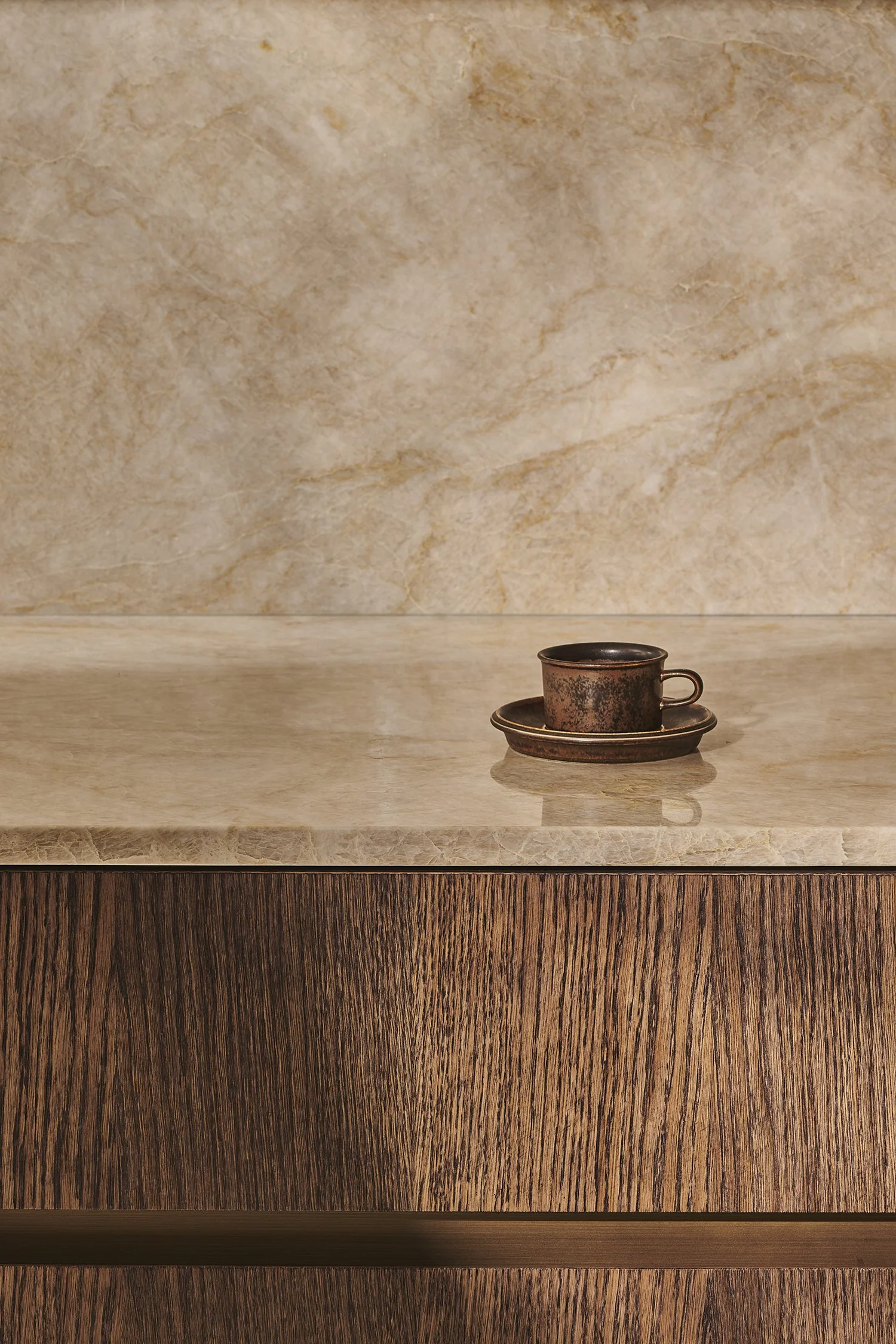 Small weathered copper cup on a matching saucer, placed on a polished beige marble countertop with a wooden drawer below.