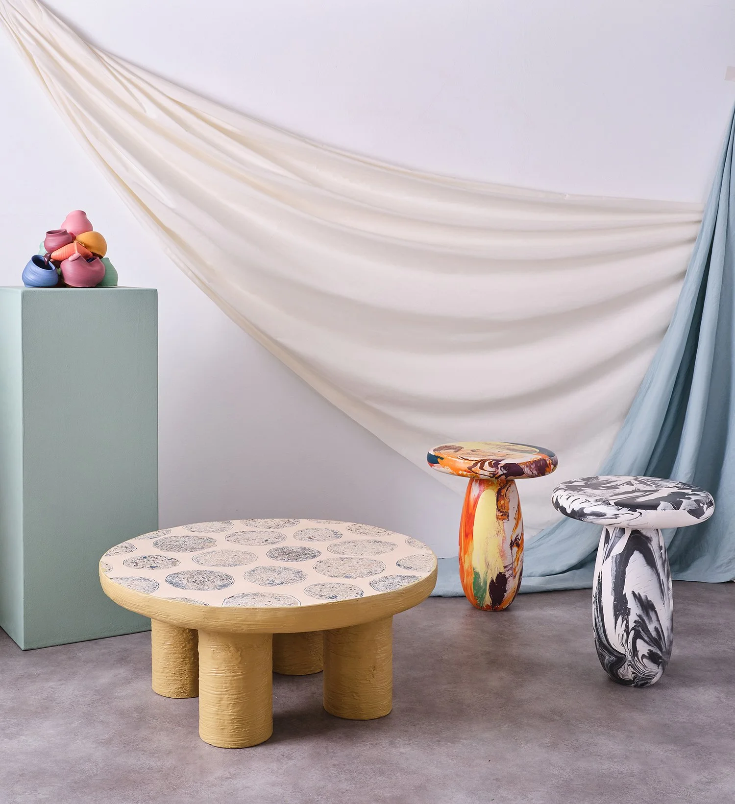 Decorative studio setup with pastel-colored draped fabric, a mint green pedestal with colorful pottery, a low beige table with circular patterns, and two marbled side tables with bright, swirling patterns.