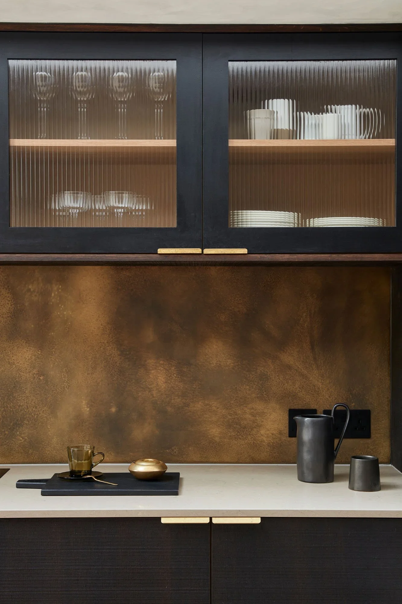 Kitchen cabinet with glass doors containing dishes and glasses, a countertop with a black tray, a gold bowl, a coffee mug, a black pitcher, and a small container, against a textured brown wall.