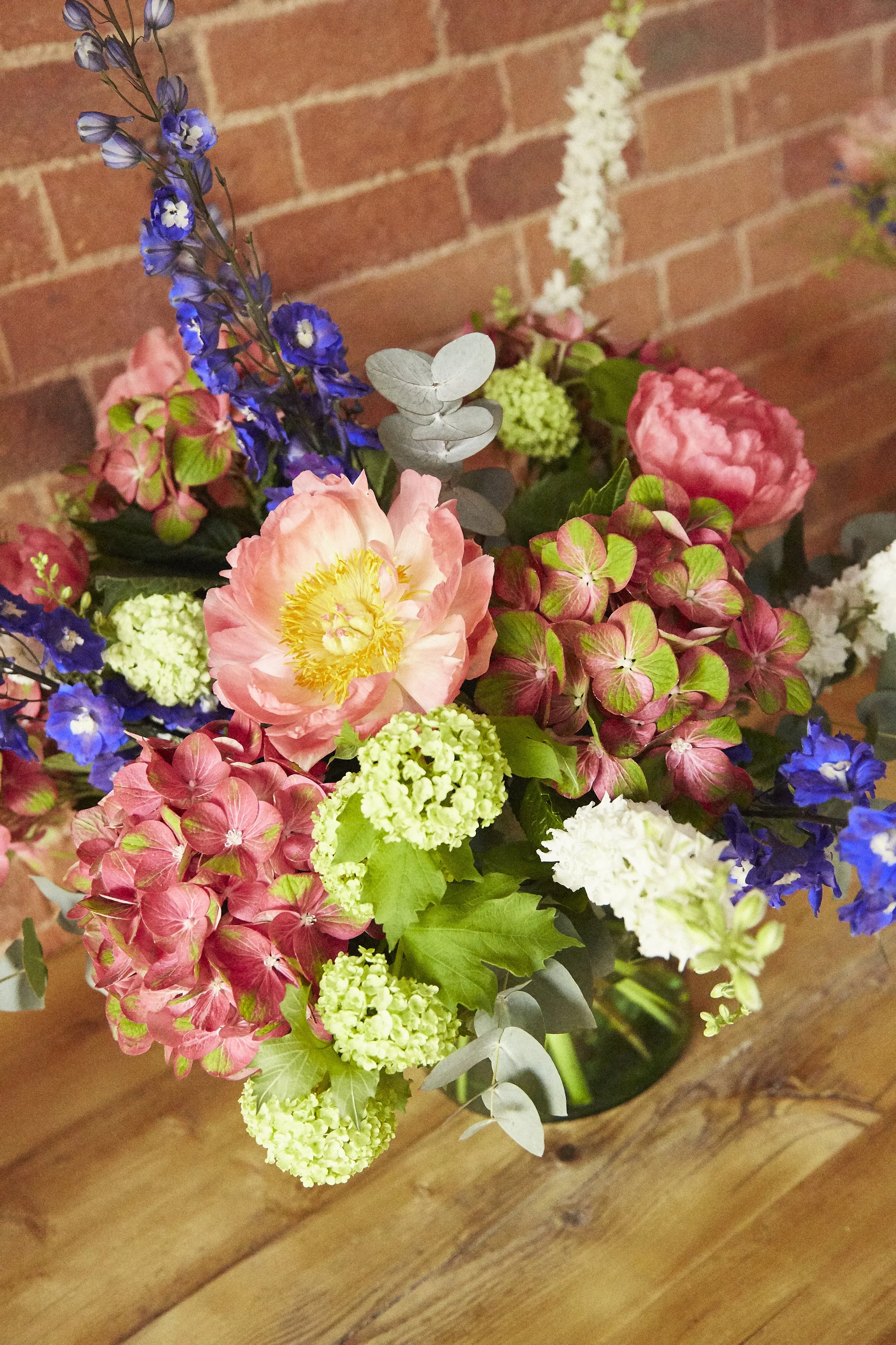 Colorful bouquet of flowers including pink peonies, purple delphiniums, green hydrangeas, and eucalyptus in a black vase on a wooden table with a brick wall background.