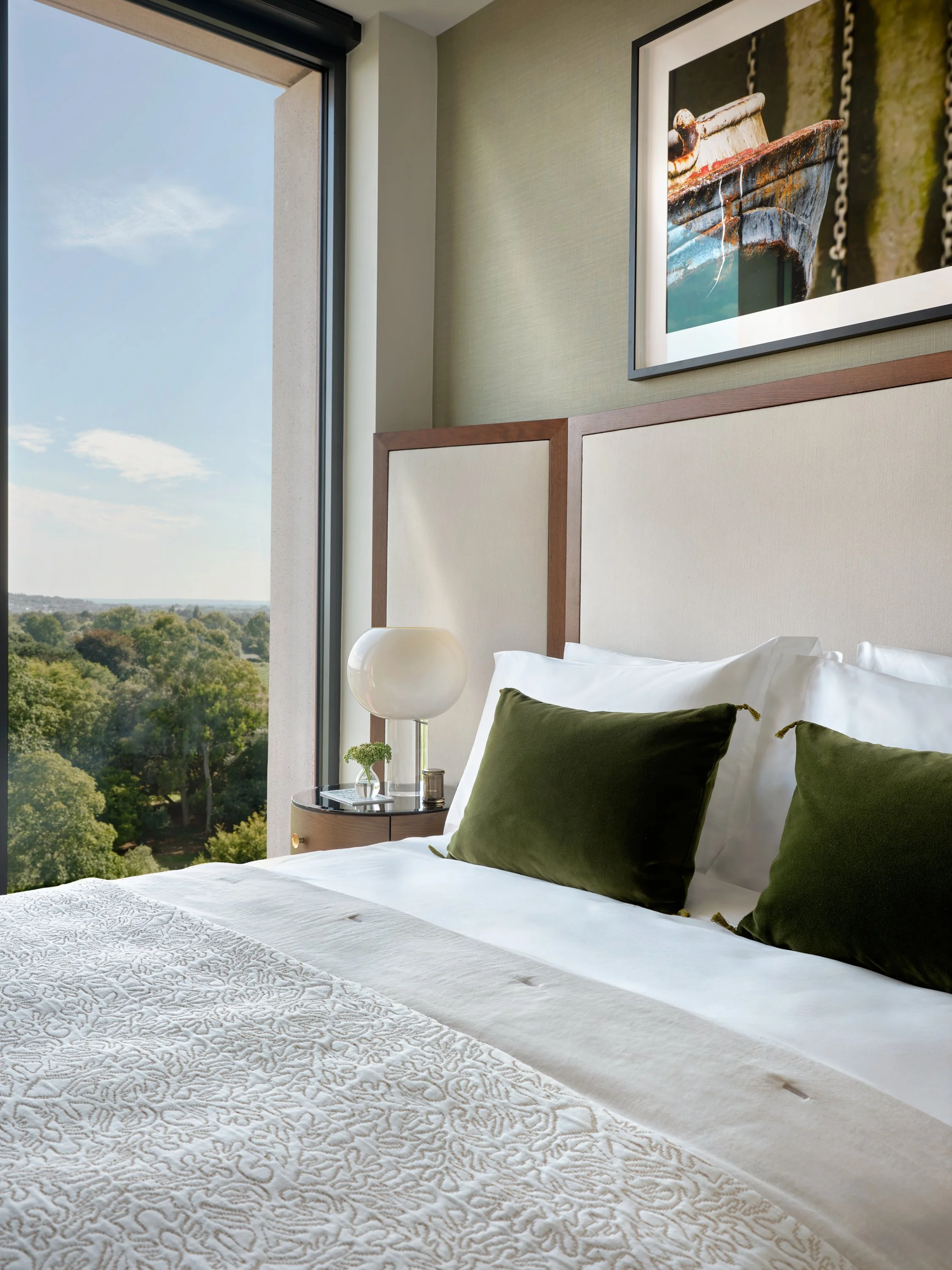 A bedroom with a large window showing a view of trees outside. The bed has white linens, green pillows, and a decorative white bedspread. There is a round bedside table with a white lamp and a small plant.