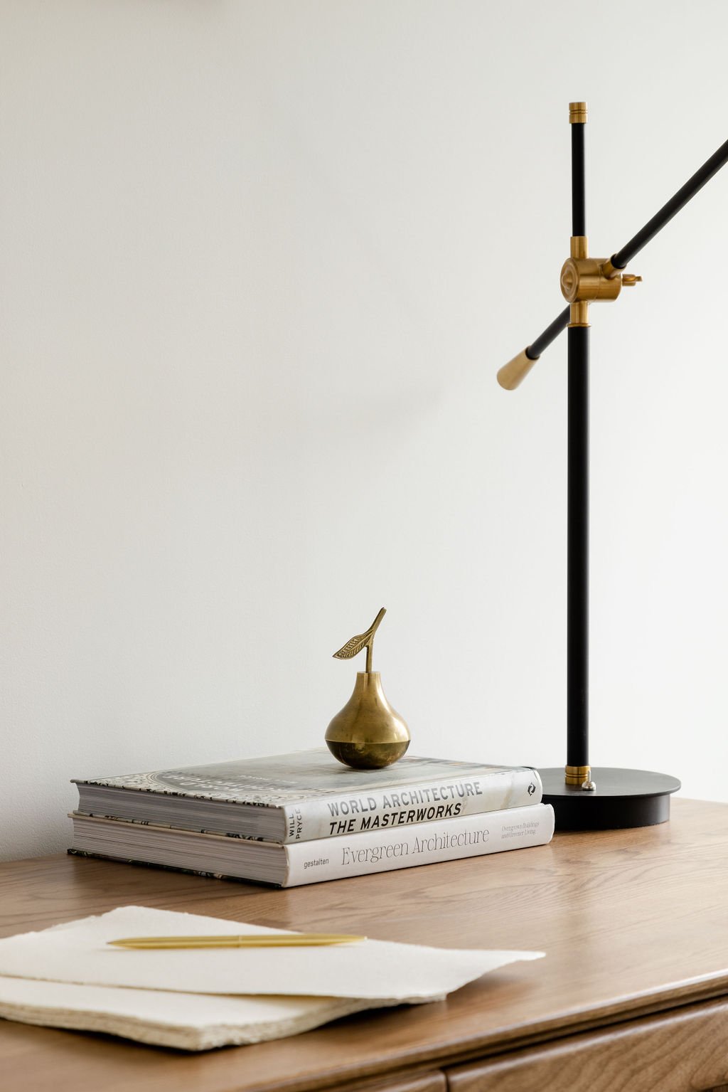 A wooden desk with books titled "World Architecture: The Masterworks" and "Evergreen Architecture," a brass vase with a leaf-shaped handle, a gold pen on a piece of paper, and a black and gold adjustable desk lamp.